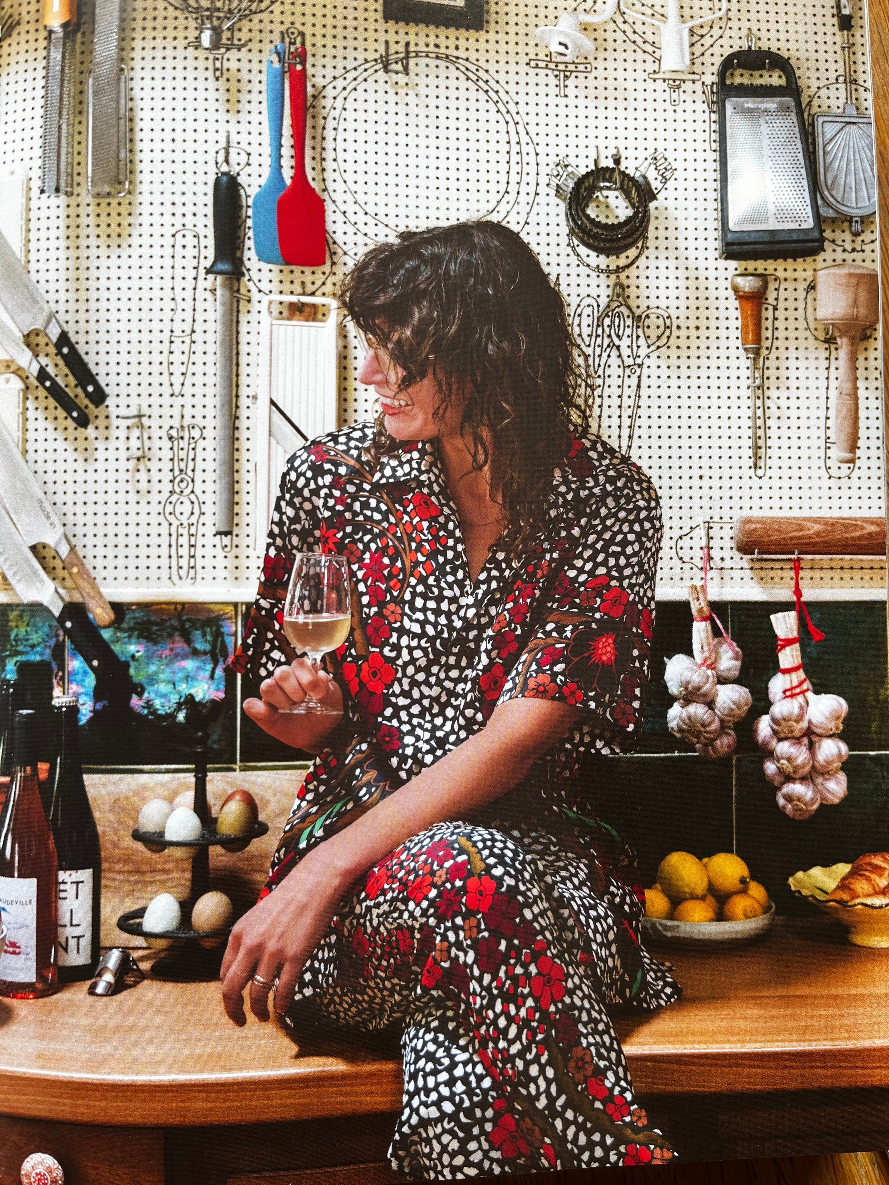 Person in floral outfit sits on kitchen counter with wine. Pegboard with utensils behind. Counter has produce and garlic.