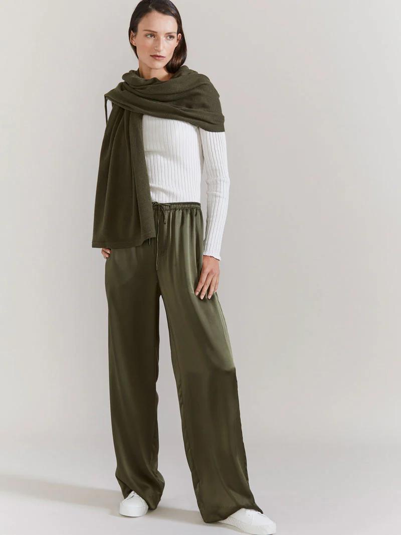 Woman in white top, olive shawl and pants, white sneakers. Standing against plain background, looking sideways with neutral expression.