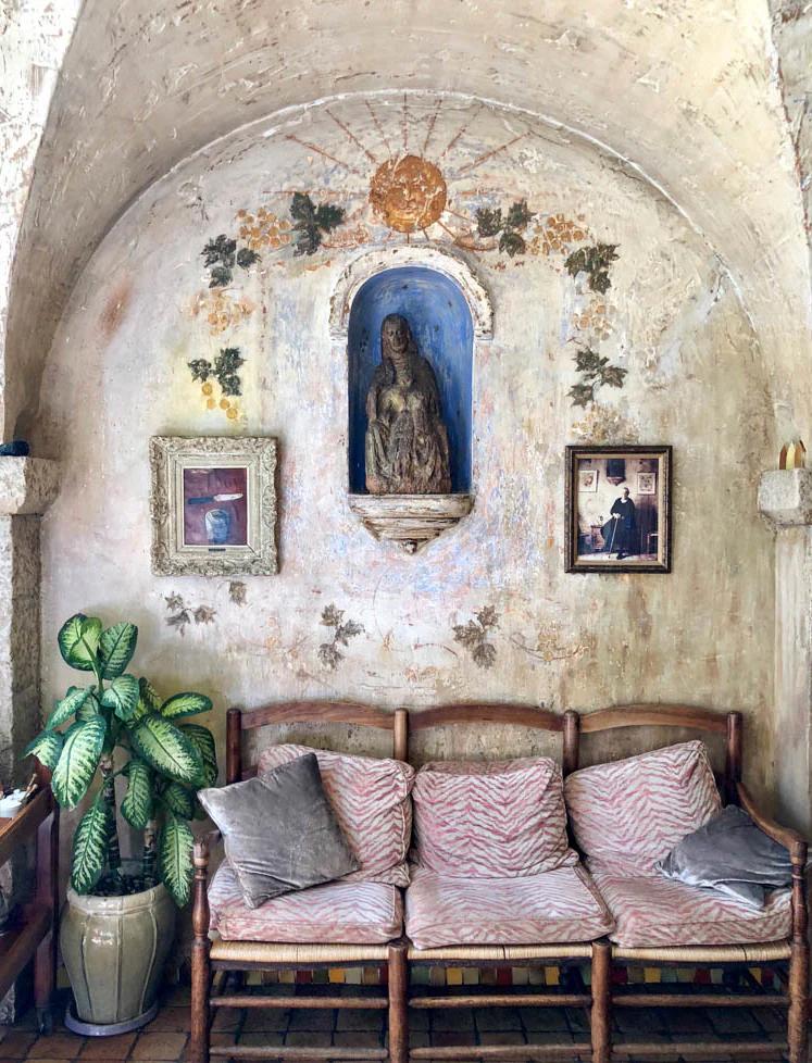 Arched alcove with Virgin Mary statue and ivy. Framed pictures on wall. Wooden bench with cushions and potted plant below.