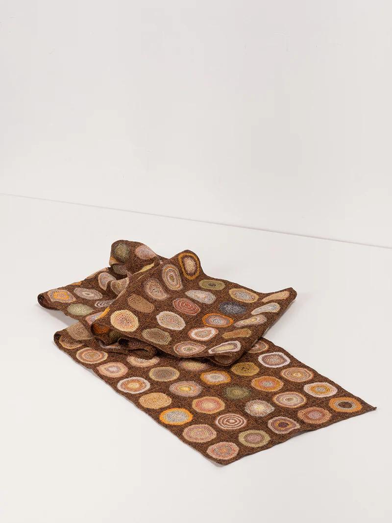 A folded brown scarf adorned with colourful crochet circles laid on a white surface. The vibrant patterns contrast with the earthy fabric, showcasing Sophie Digard’s intricate design.