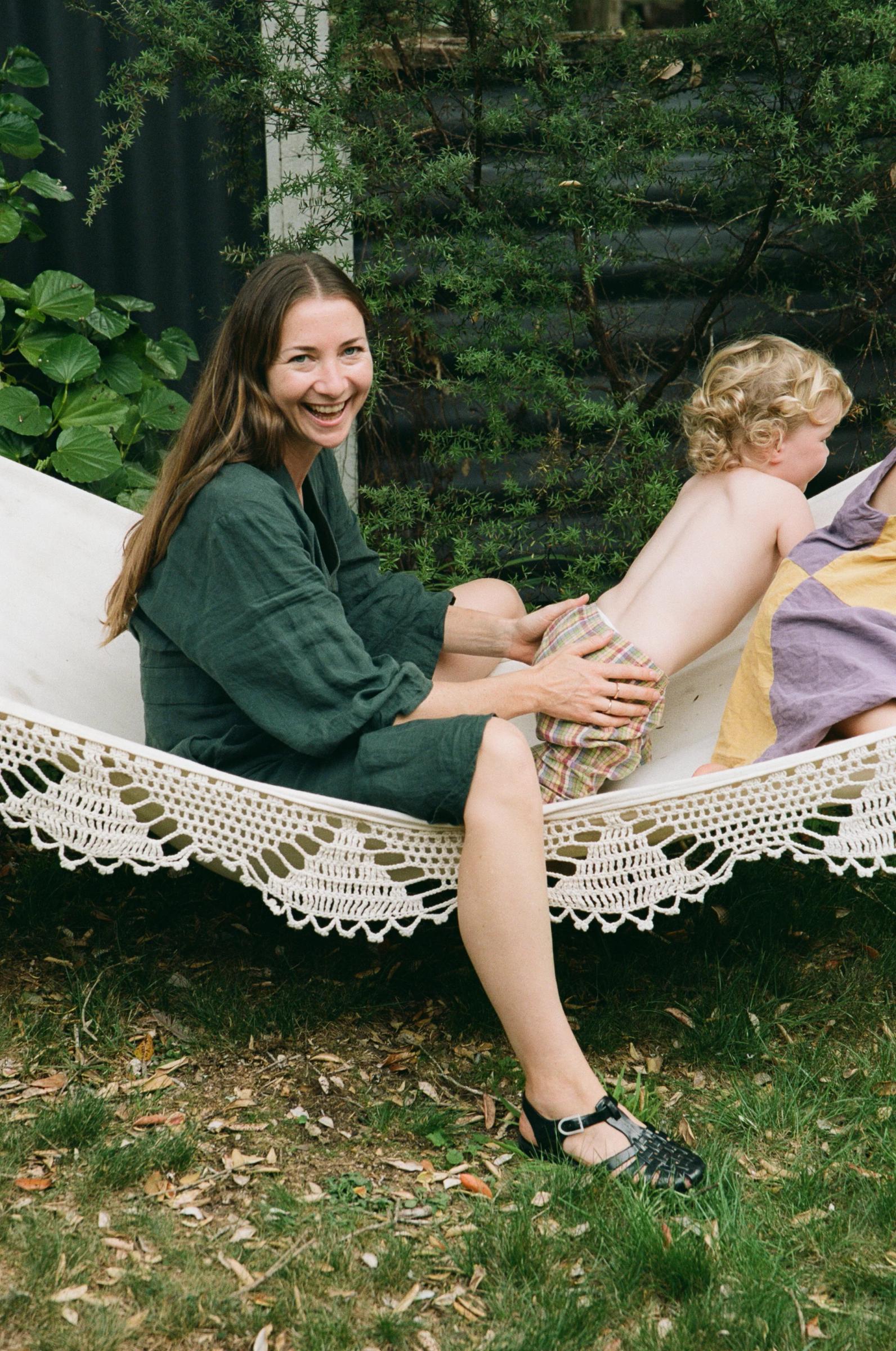 Woman in green dress on white hammock holding blonde child. Green foliage and dark building in background.