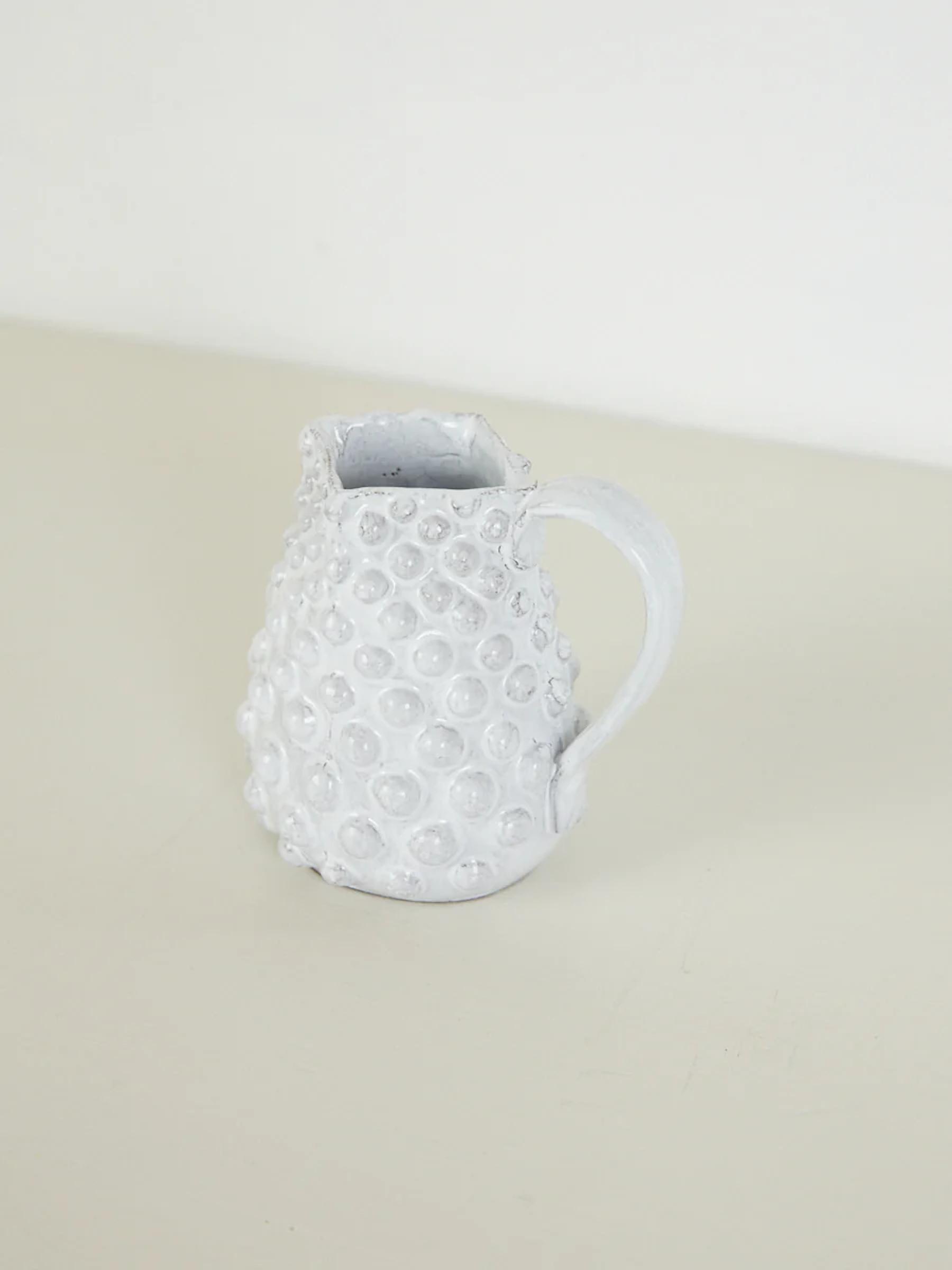 Small white ceramic pitcher with textured surface. Circular bumps, handle. Light-colored surface.