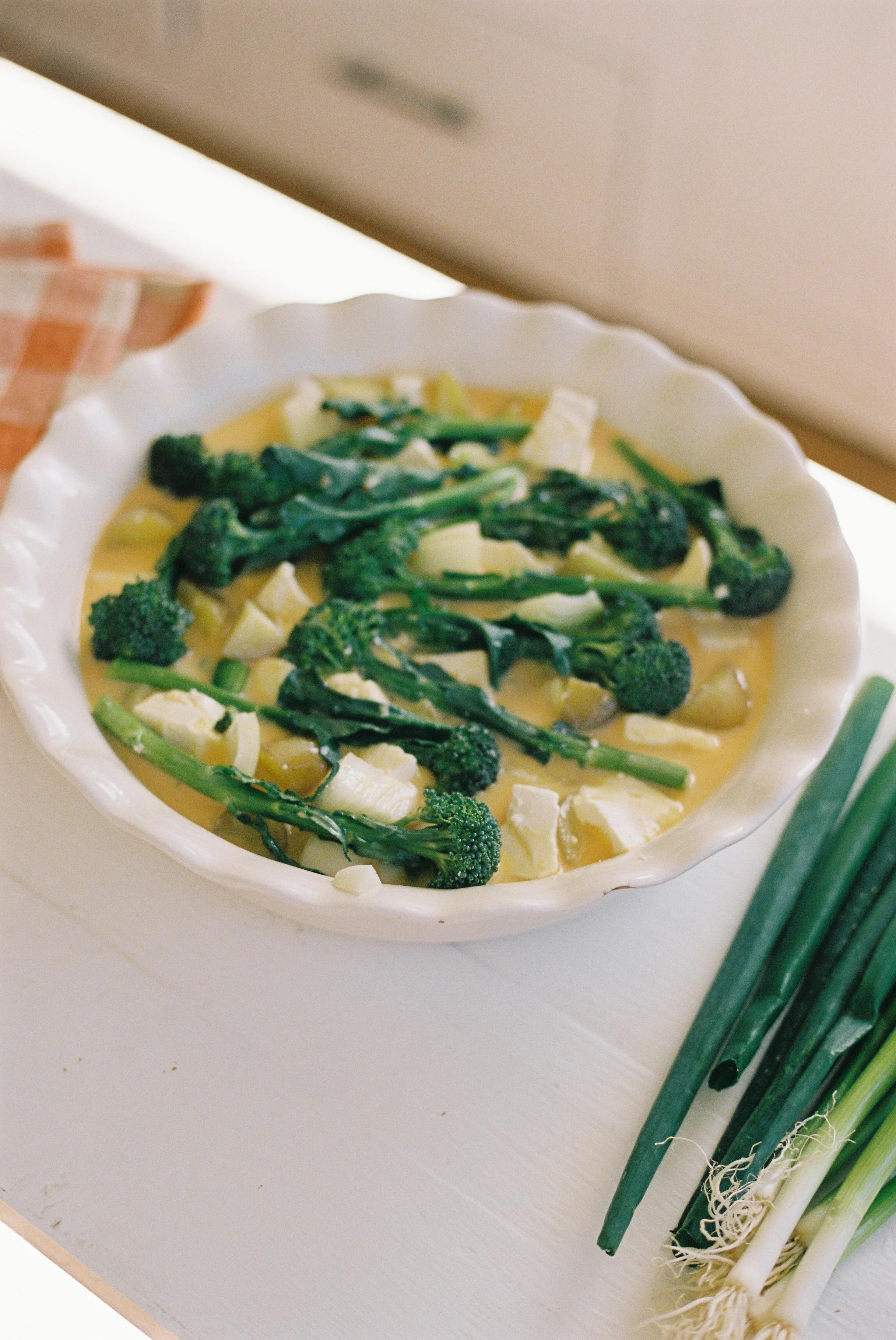 Broccoli, onion, and brie quiche in a white scalloped dish. Set on a countertop with gingham napkin and fresh green onions for a springtime meal.