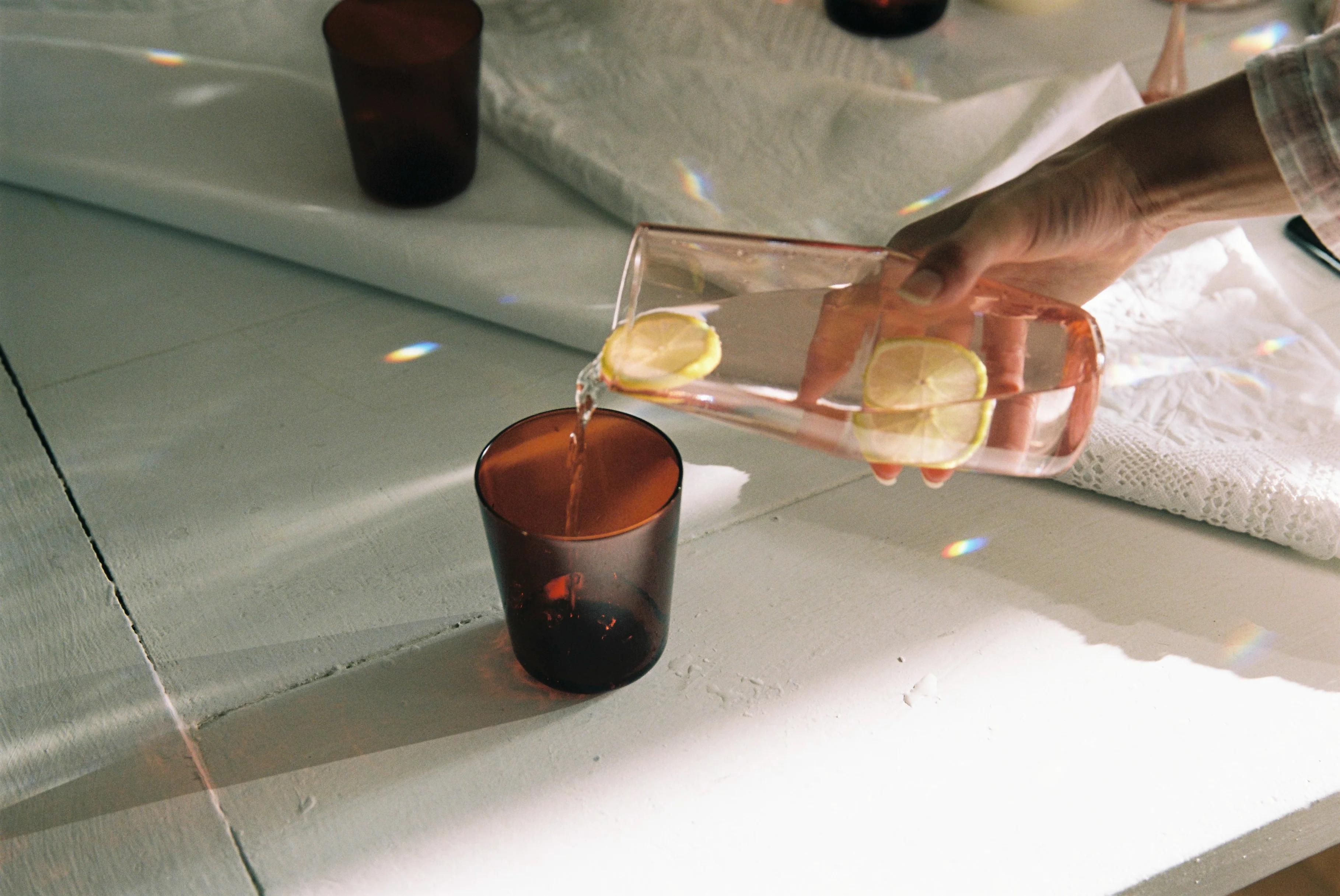 A hand pours water with lemon slices from a clear R+D.LAB carafe into an amber tumbler on a white surface. Sunlight creates prism reflections with blurred dark glasses in the background.