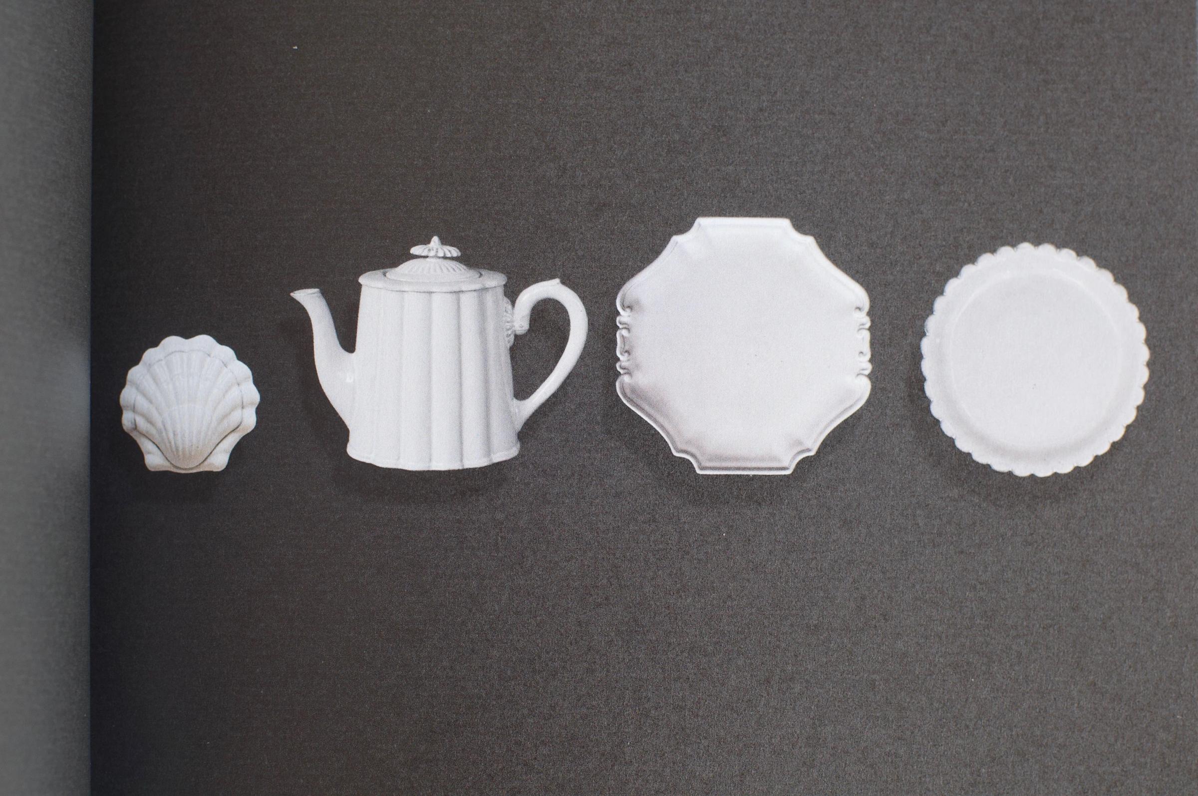Four white porcelain items on a dark background: a shell-shaped dish, teapot, square plate with scalloped edges, and a textured round plate.