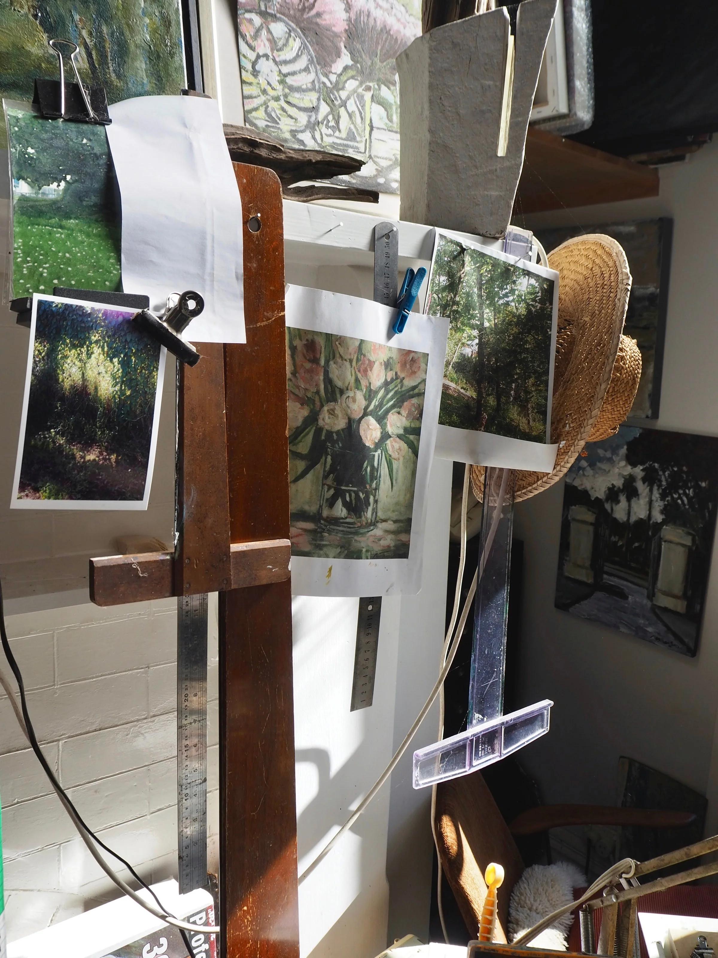 Cluttered studio with Max Thomson's paintings and photos. Straw hat on wall. Brushes, canvases scattered in natural light.