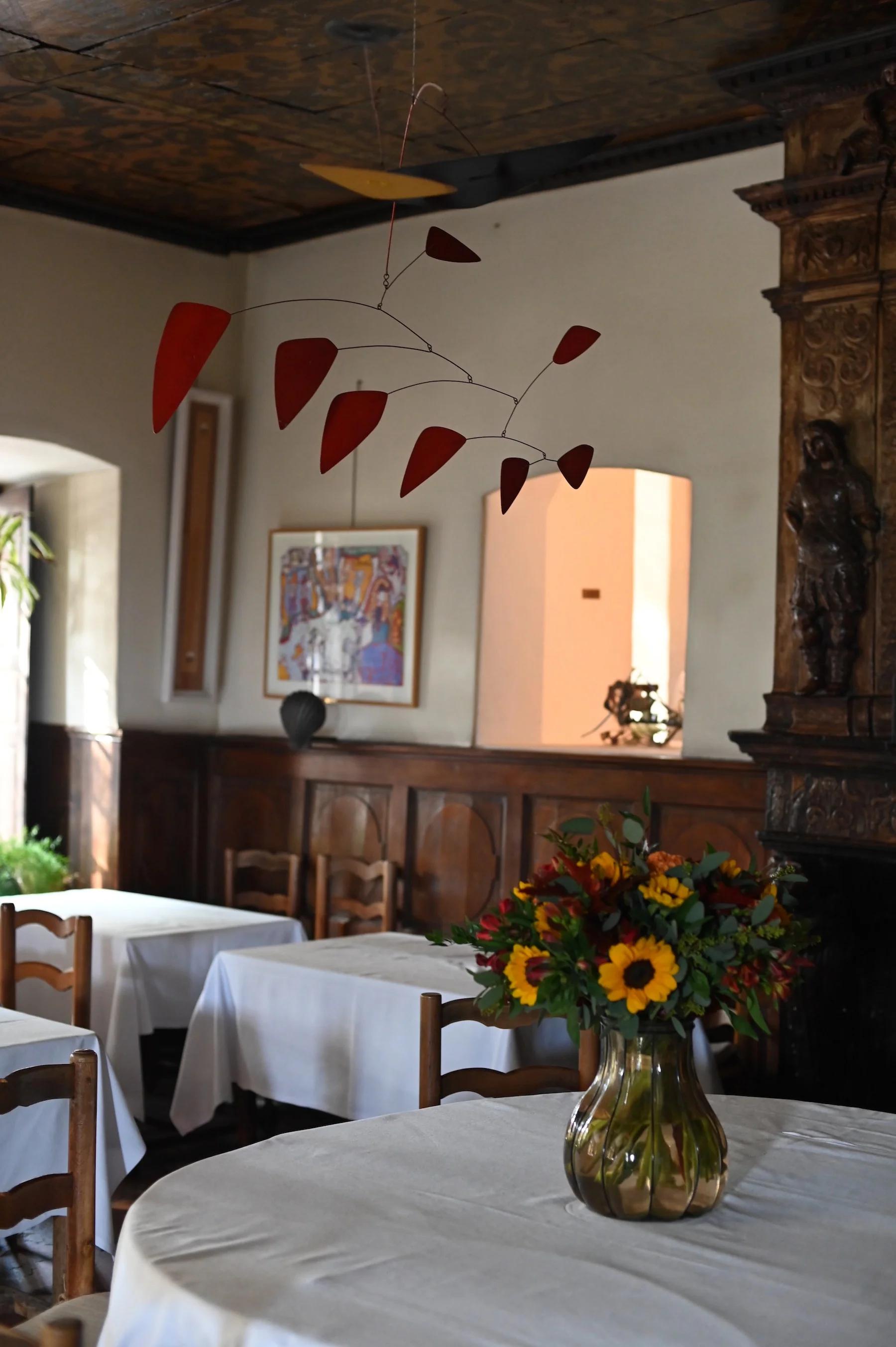 Cozy restaurant with white-clothed tables, wooden chairs, and fireplace. Colourful artwork and modern red mobile decorate the space.