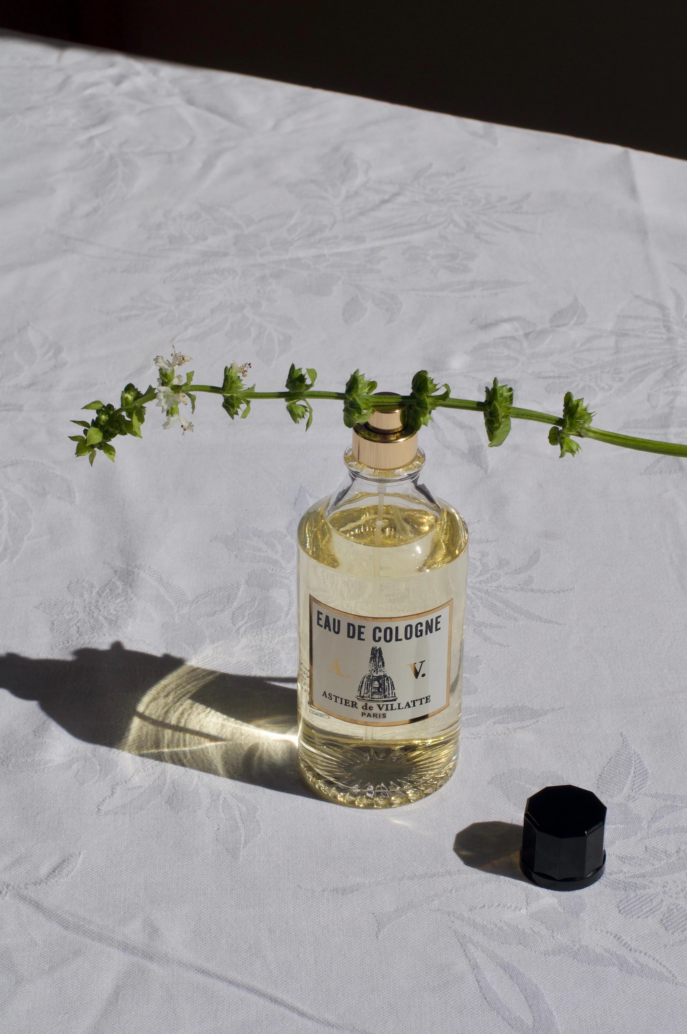 Glass "Eau de Cologne" bottle on patterned tablecloth. Fresh greens and flowers on top. Black cap beside bottle.