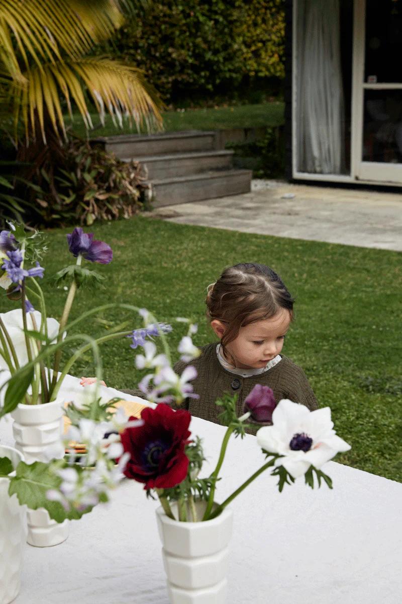 A child with dark braided hair stands in a garden, looking down. Colourful flowers are arranged nearby for Rachel Carley’s dinner party, with greenery and a building entrance in the background.