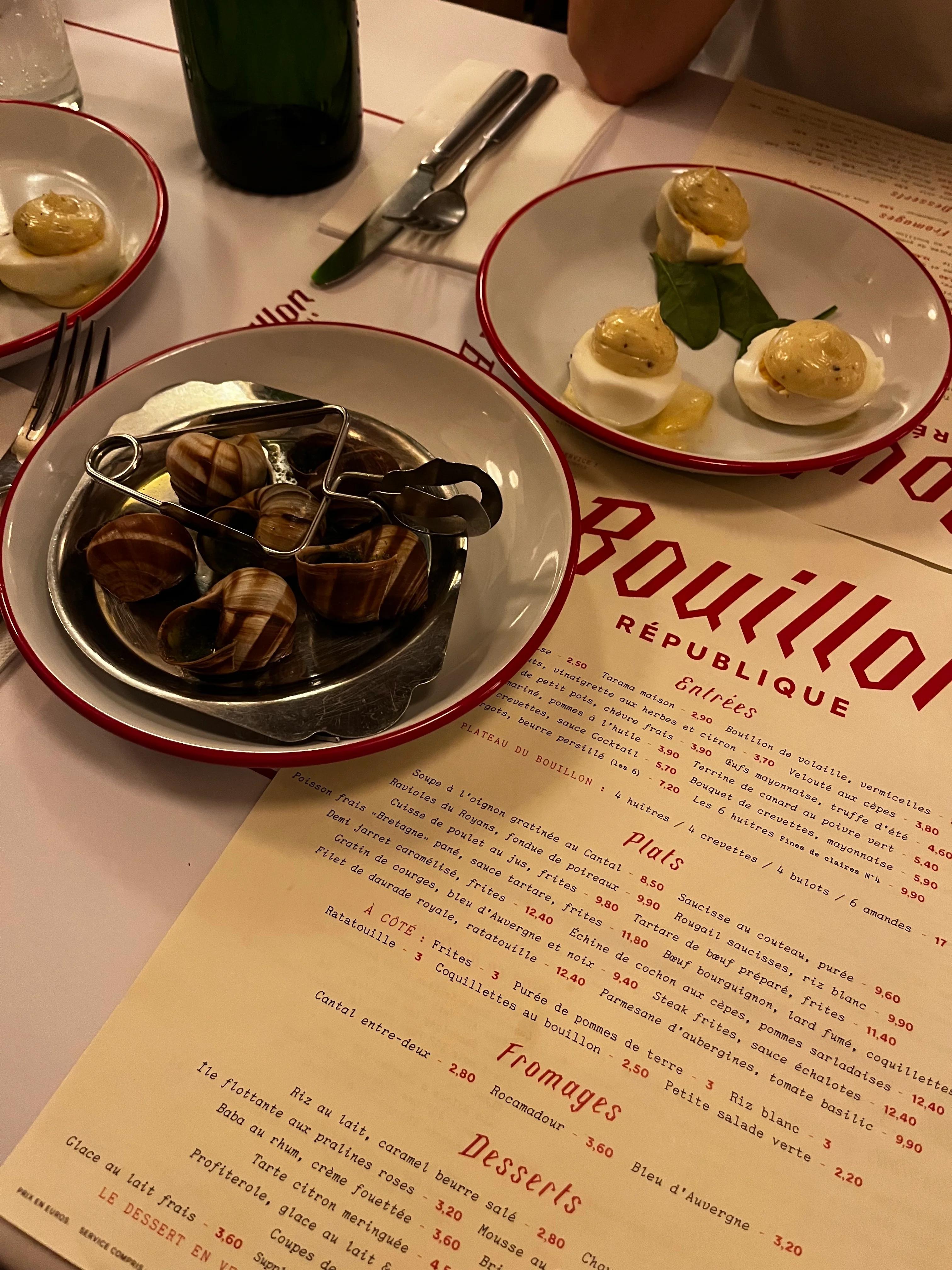 Restaurant table with deviled eggs and escargot. Utensils and "Bouillon République" menu. Green bottle partially visible.