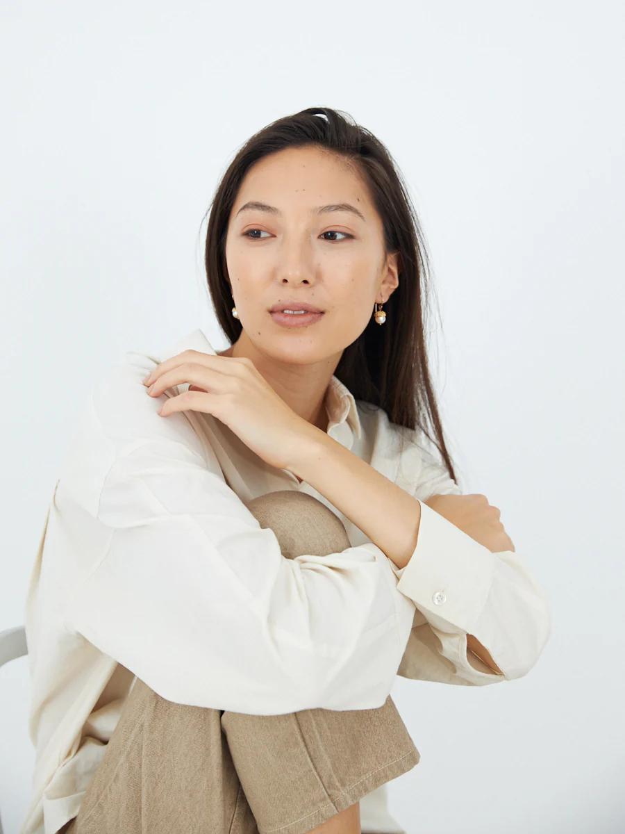 Person with long brown hair in cream blouse and beige pants sits looking to side. White background.