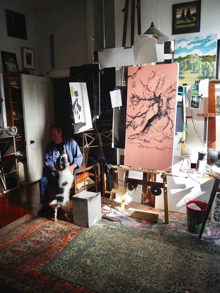 Thomson petting black and white dog in art-filled studio. Paintings, easels, and supplies surround them. Sunlight casts shadows on floor and rugs.