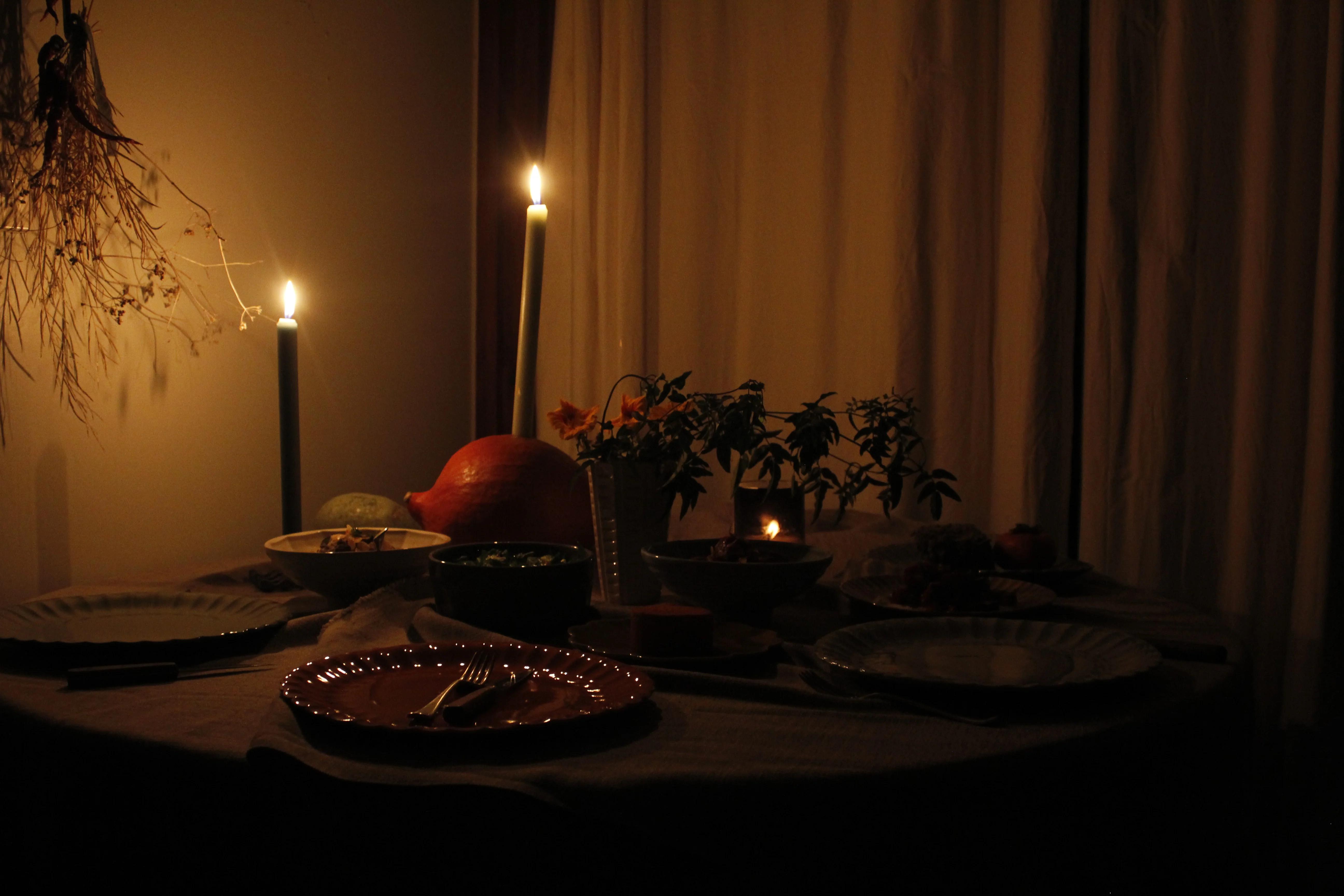 A dimly lit table features candles, Palestinian dishes, and a potted plant, with dried plants and curtains adorning the background.