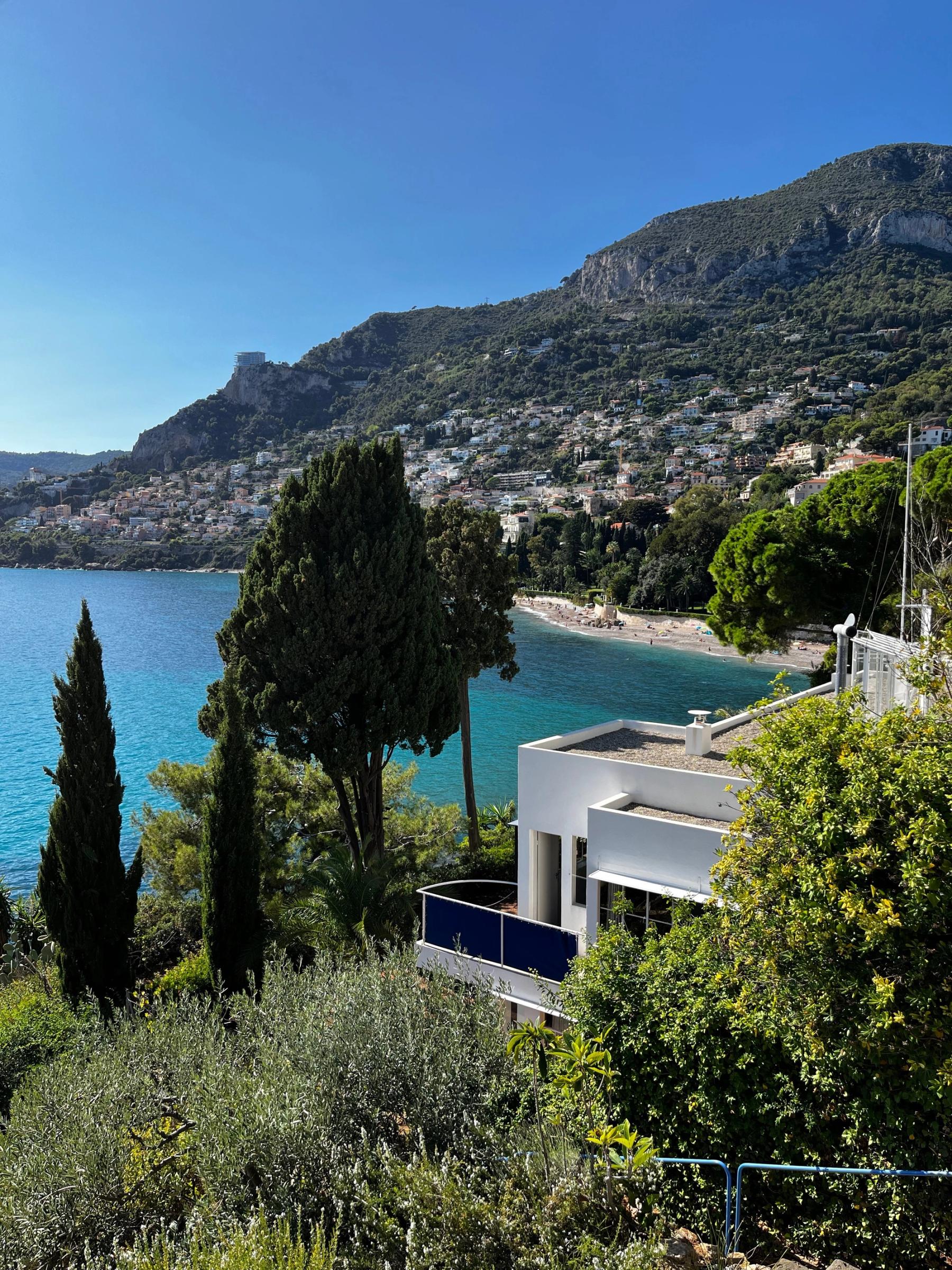 Coastal view with beach, greenery, and white buildings. Modern white house with blue balcony in foreground. Hills in background.