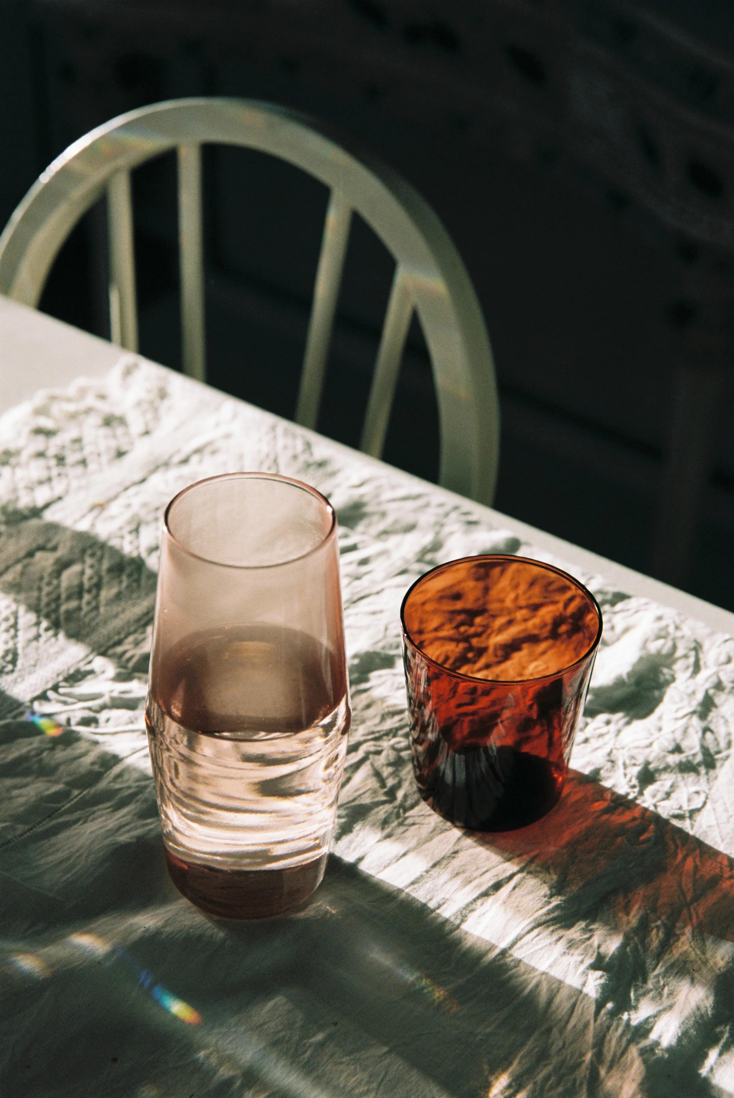 A white table with a patterned tablecloth features two glasses: one tall and transparent, the other shorter with an amber hue. A white chair with vertical slats is behind the table.