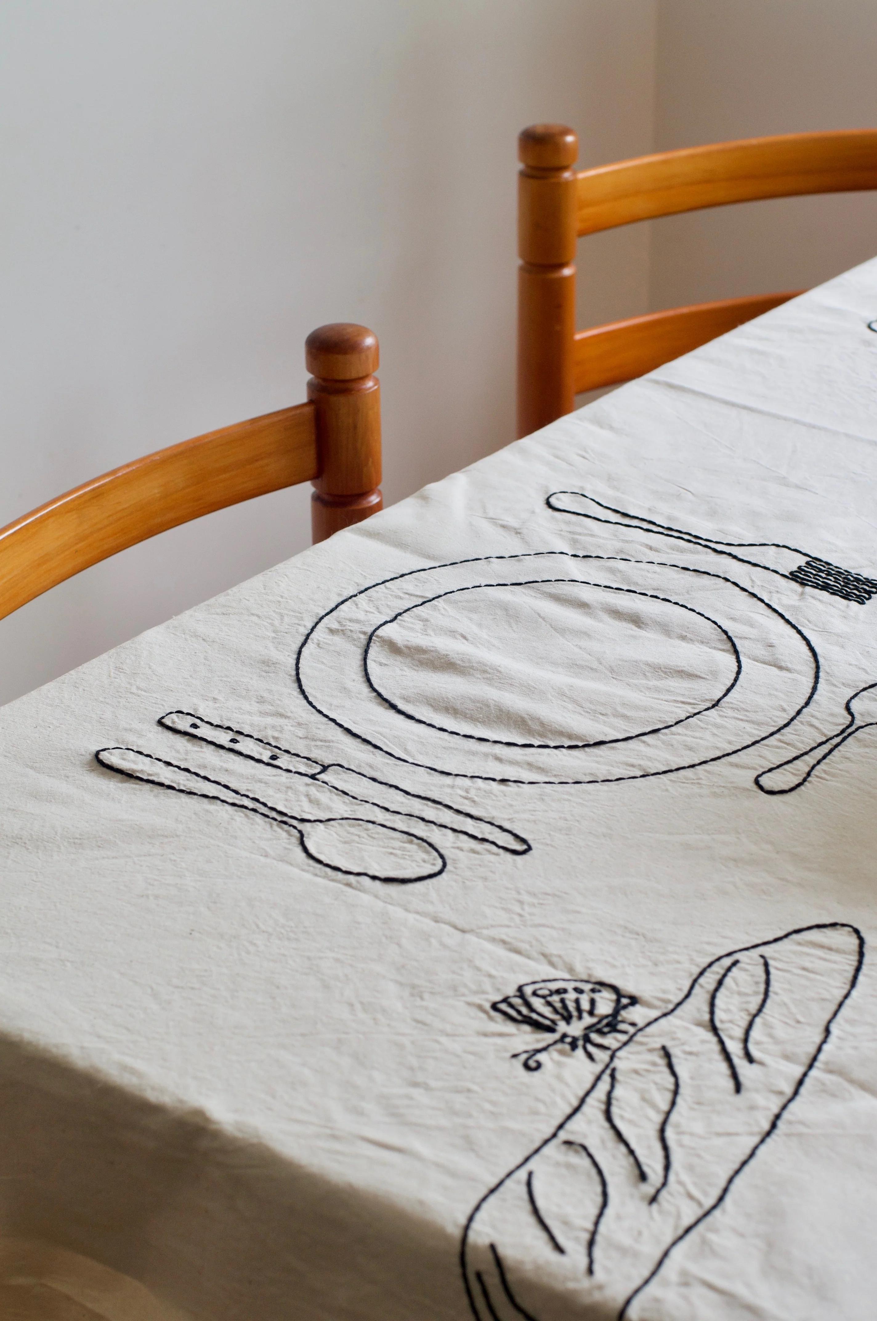 Dining table with embroidered tablecloth showing plate and cutlery outlines. Two wooden chairs flank table. Bare, neutral background.