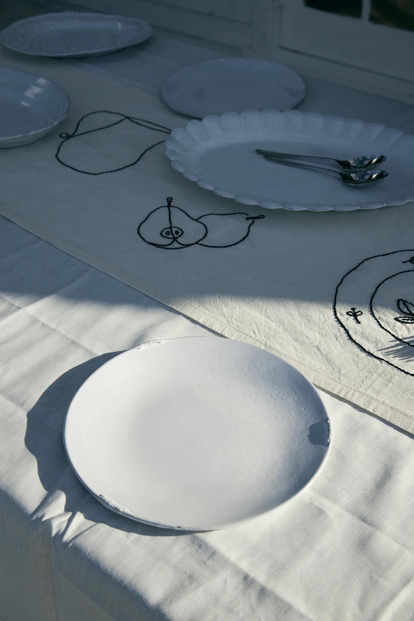 Table set with white plates and spoons. Tablecloth with minimalist pear drawings. Natural light casts soft shadows.