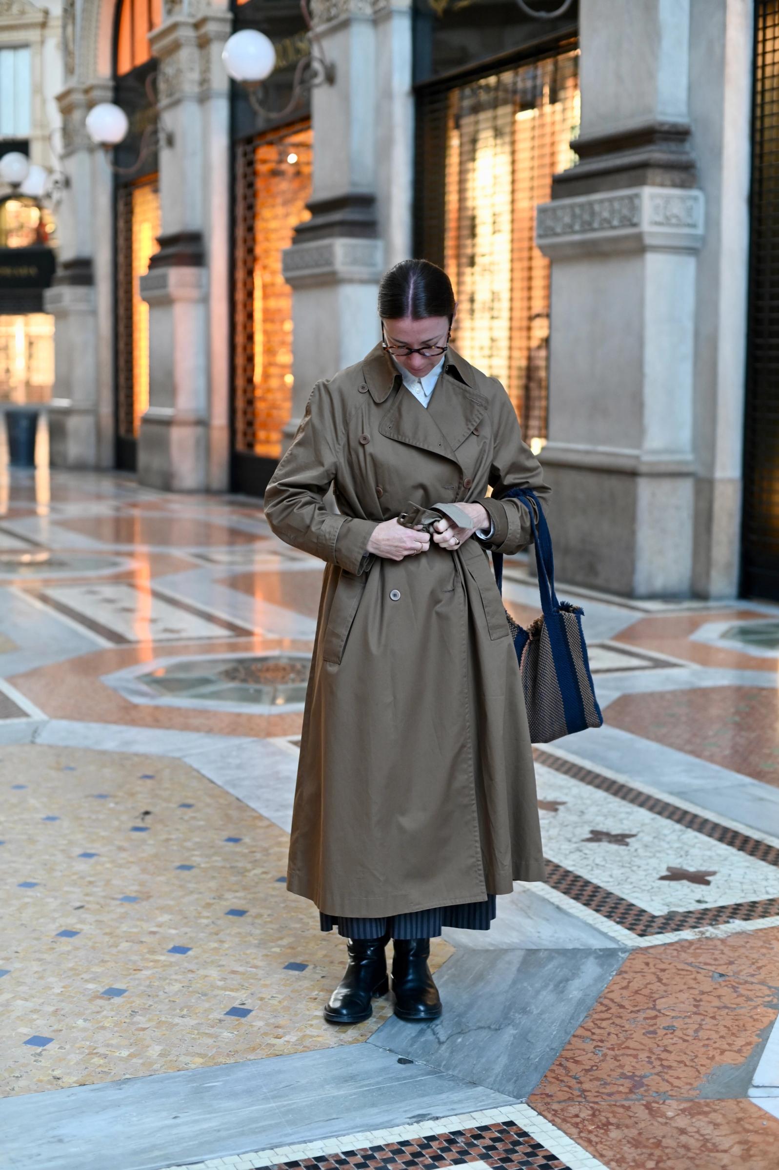 women in halls ties her trench coat up at the waist while holding a tote bag