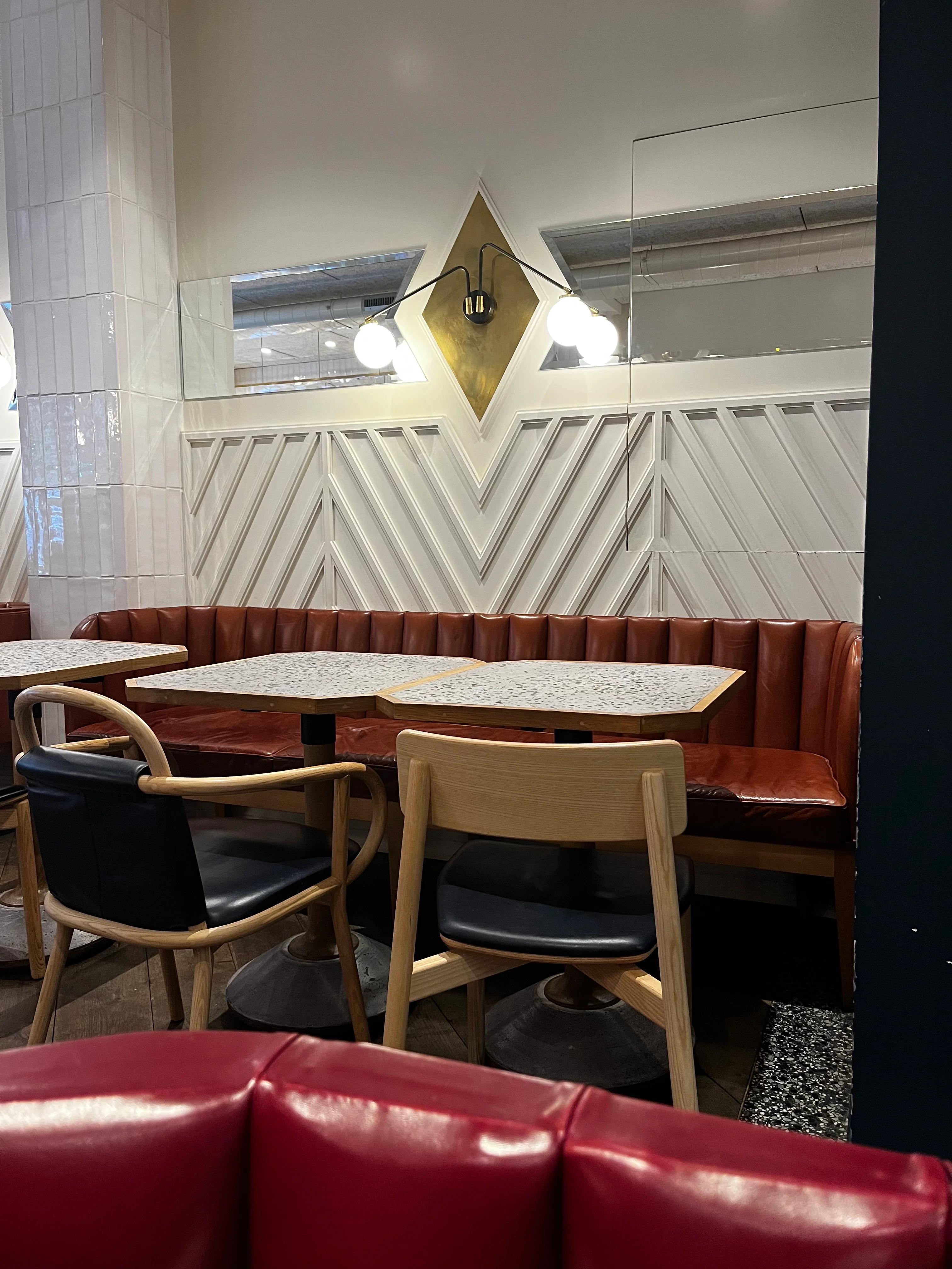 Cafe interior with red cushioned bench seating. White patterned wall with geometric decor. Marble-topped tables and mixed chairs. Modern light fixtures.