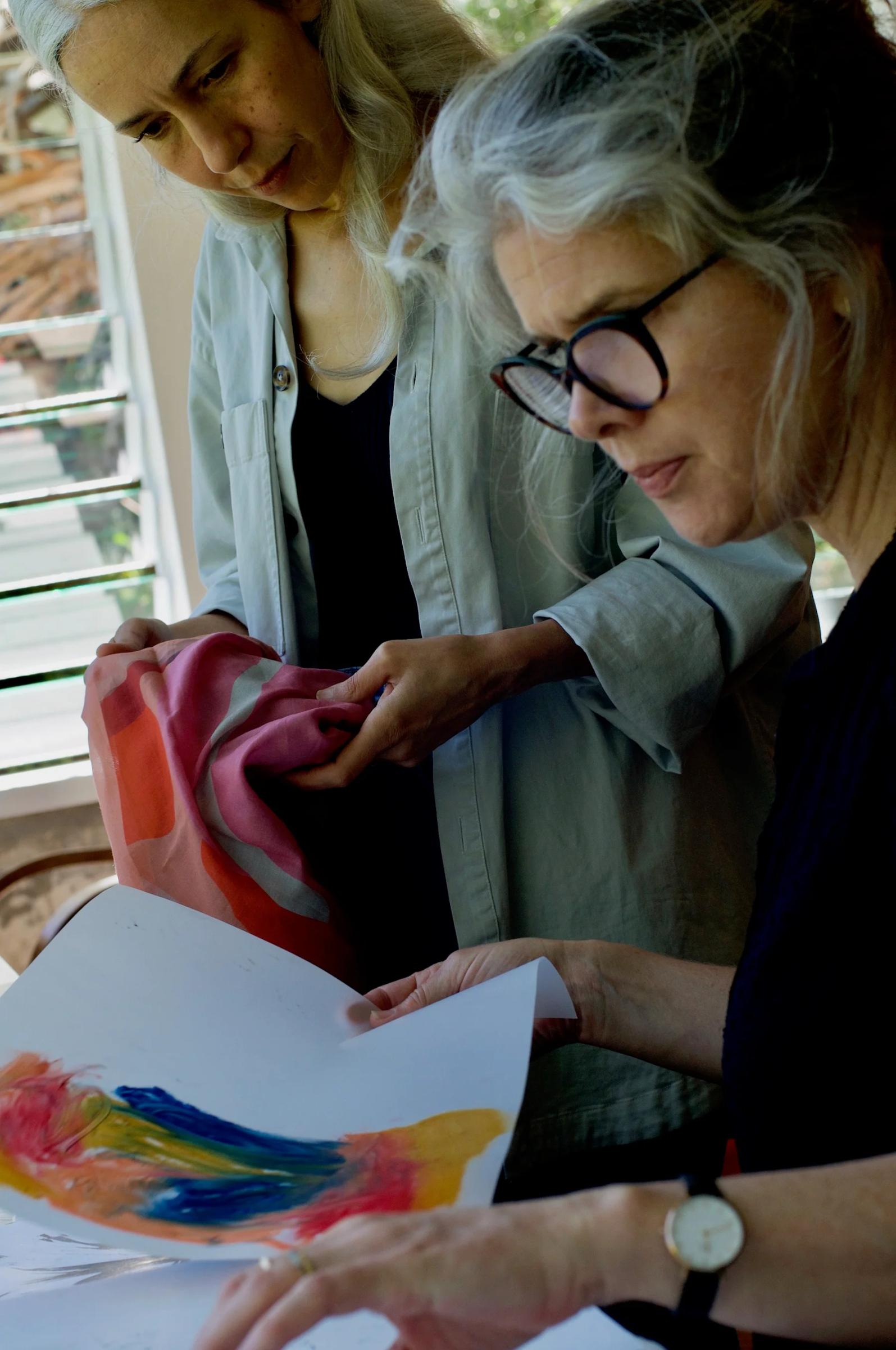 Two people examining colourful paper. One wearing glasses, other holding vibrant cloth. Indoor setting.