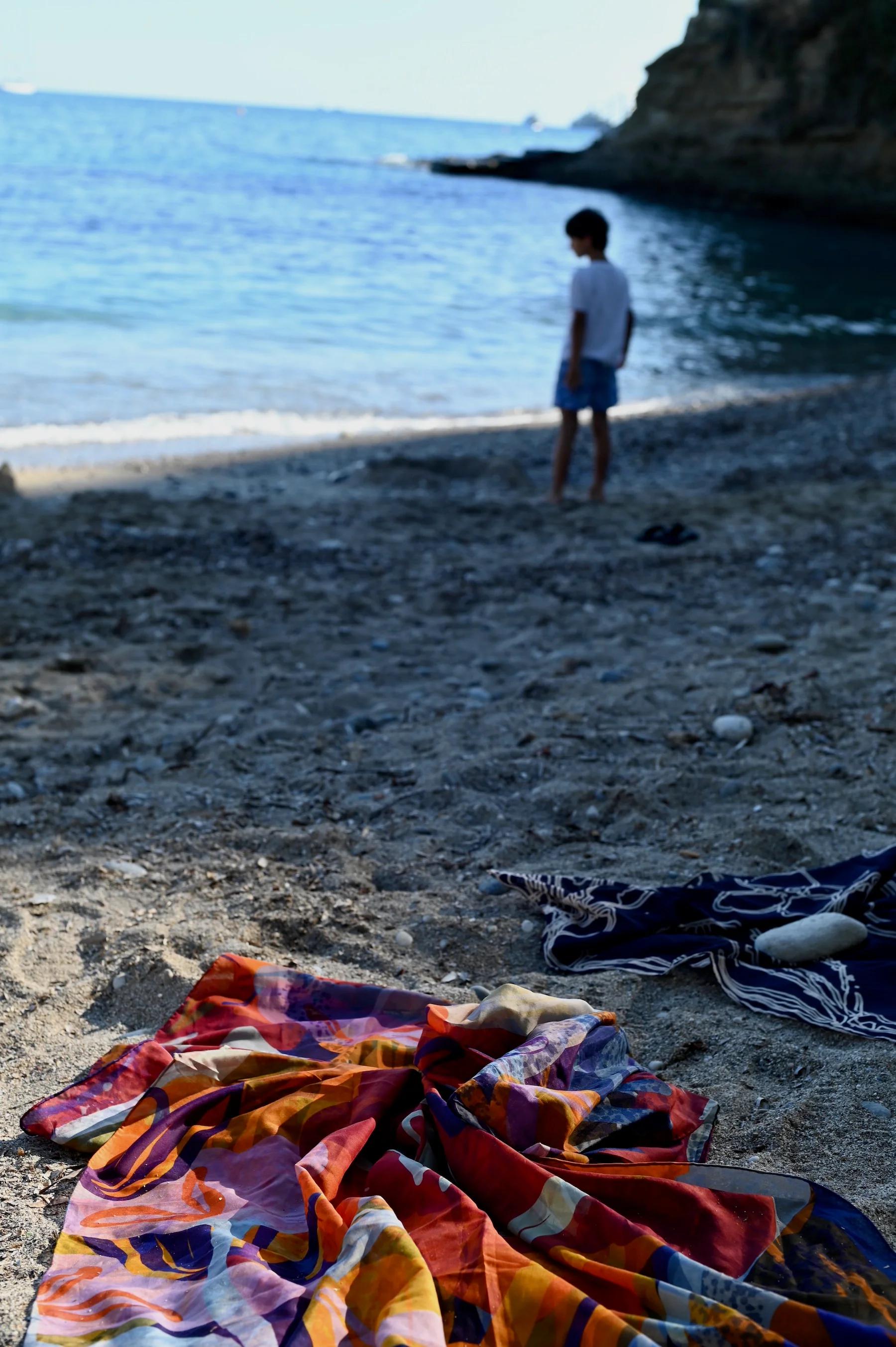 Sandy beach with colourful towels in foreground. Person in shorts and white shirt at water's edge in background. Curved shoreline, rocks and trees in distance.