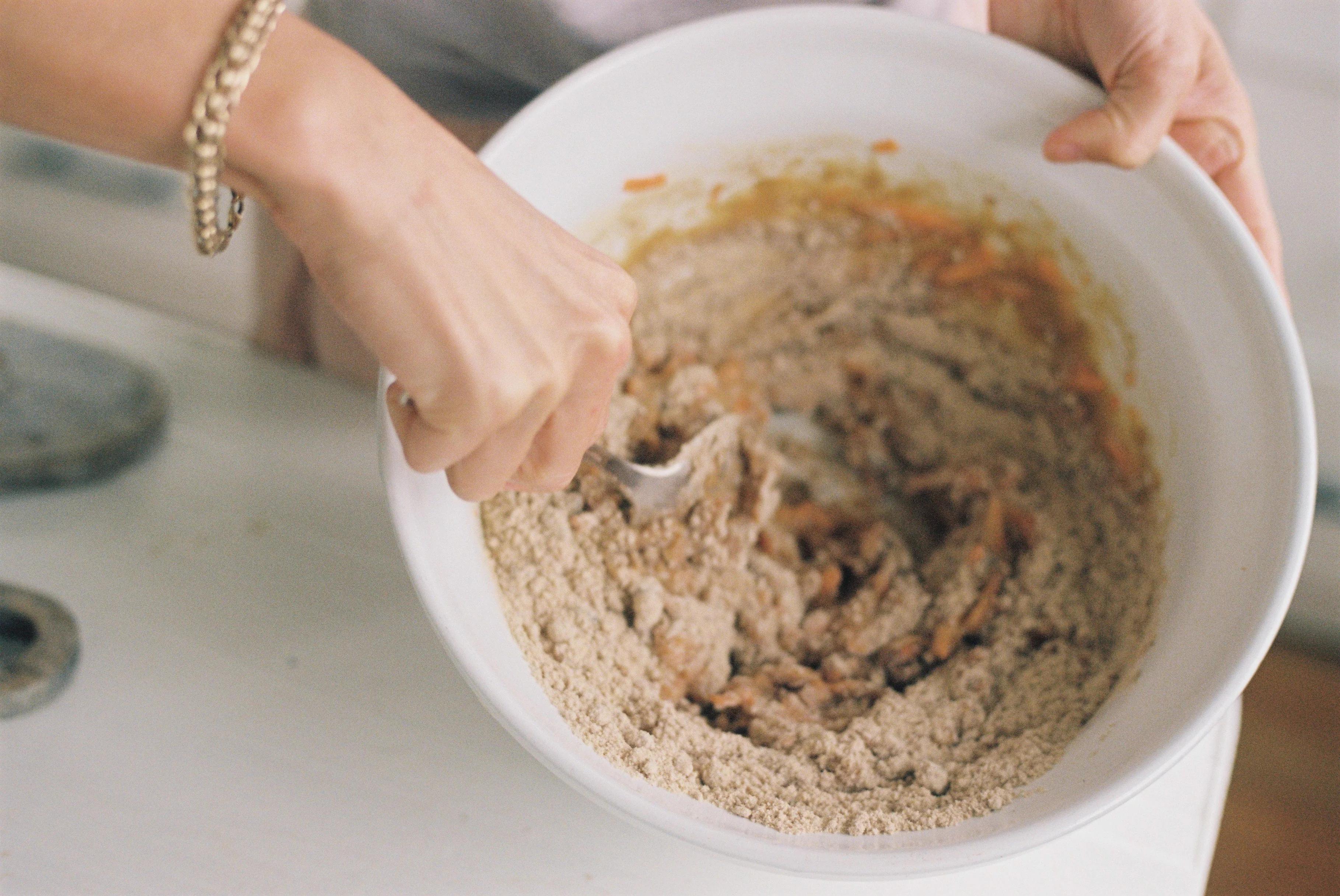 Person mixing ingredients in white bowl. Wearing bracelet. Cake kit components visible.