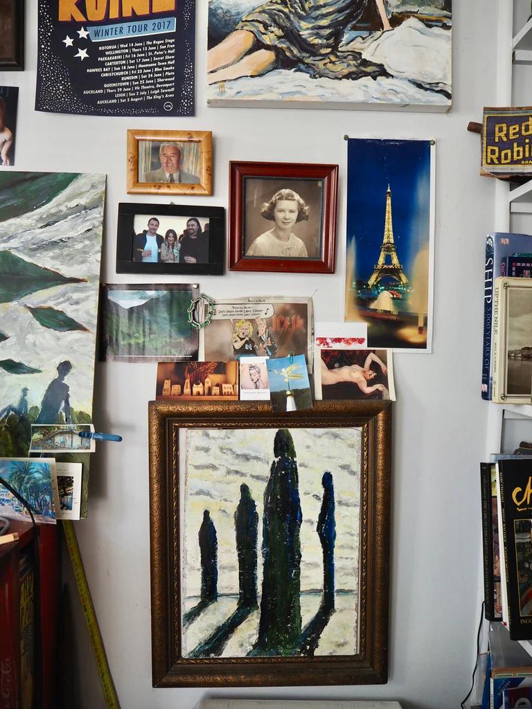 Wall covered in diverse framed art, photos, and posters. Features woman's portrait, Thomson's elongated figures painting, and lit Eiffel Tower image.