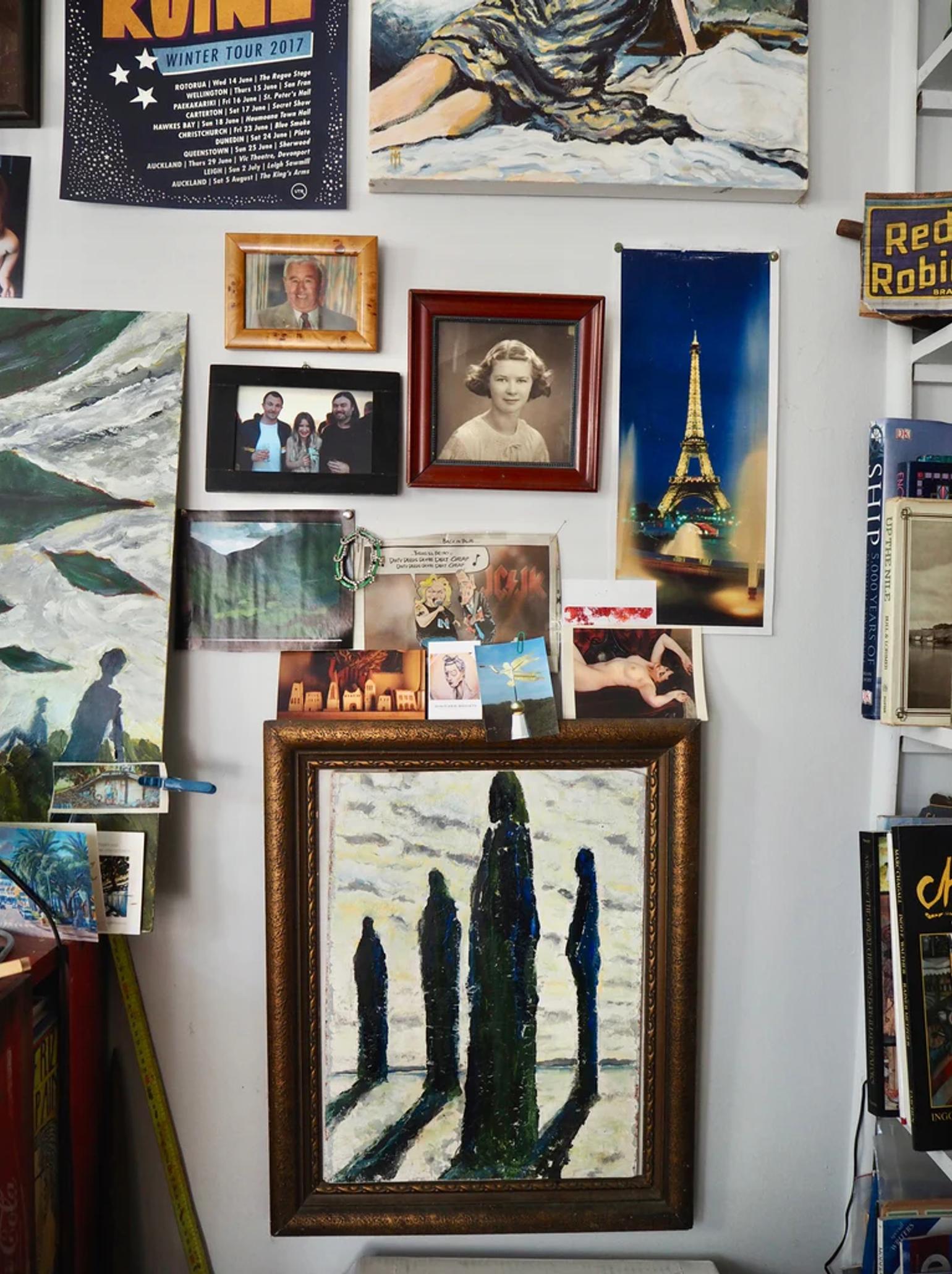 Wall covered in diverse framed art, photos, and posters. Features woman's portrait, Thomson's elongated figures painting, and lit Eiffel Tower image.