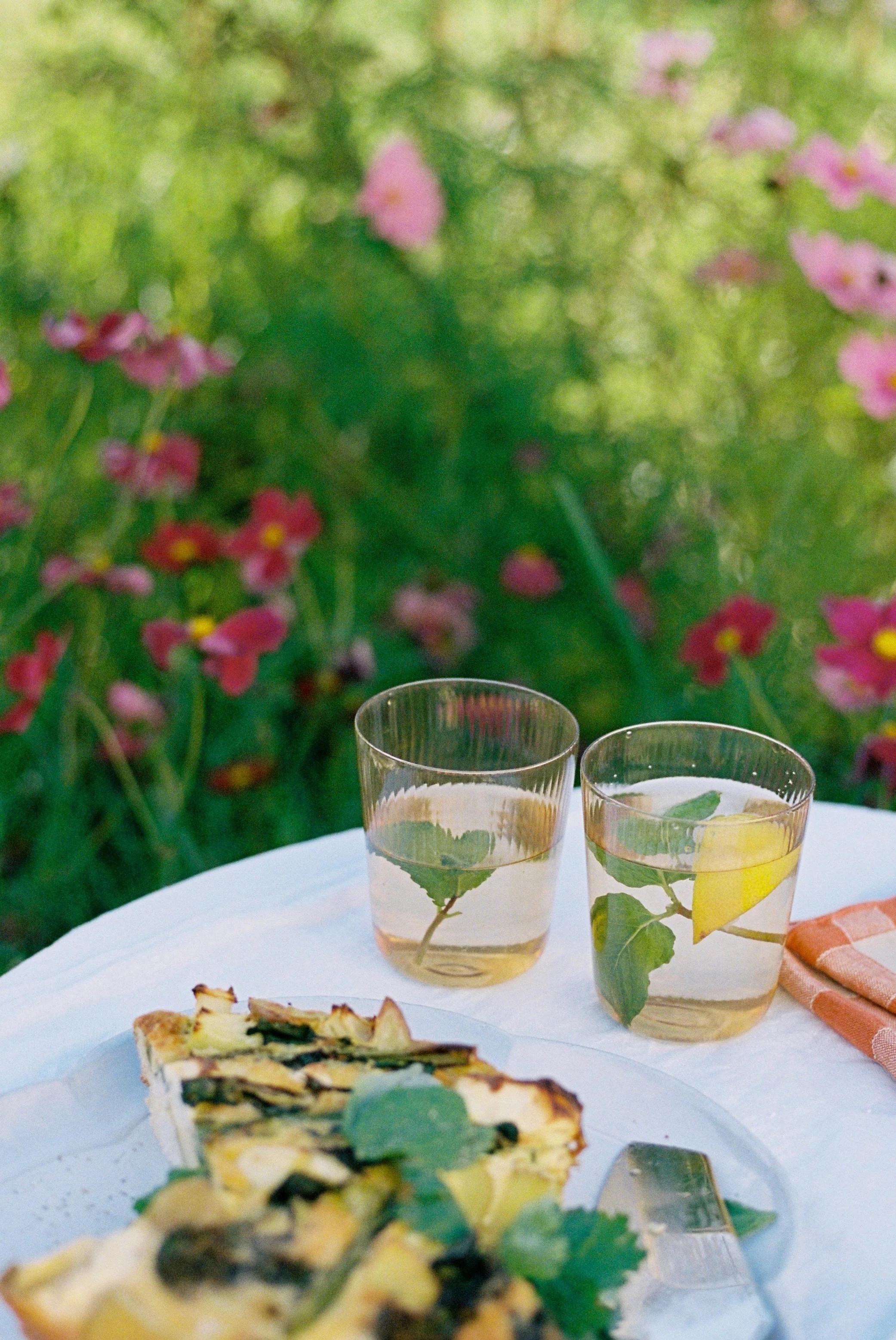 Garden table with two lemon and mint-garnished drinks. Plate of green frittata slices nearby. Colourful flowers and greenery create vibrant, serene backdrop.