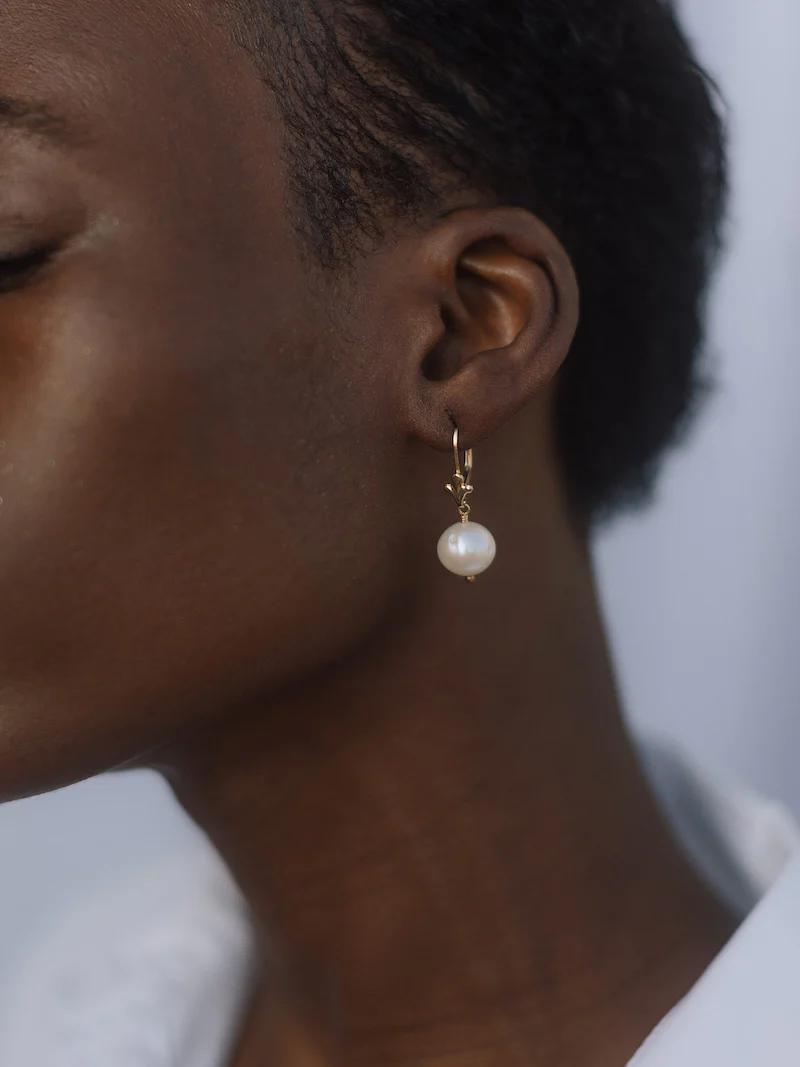 Side profile of person wearing gold earring with dangling pearl. Short dark hair, white garment.