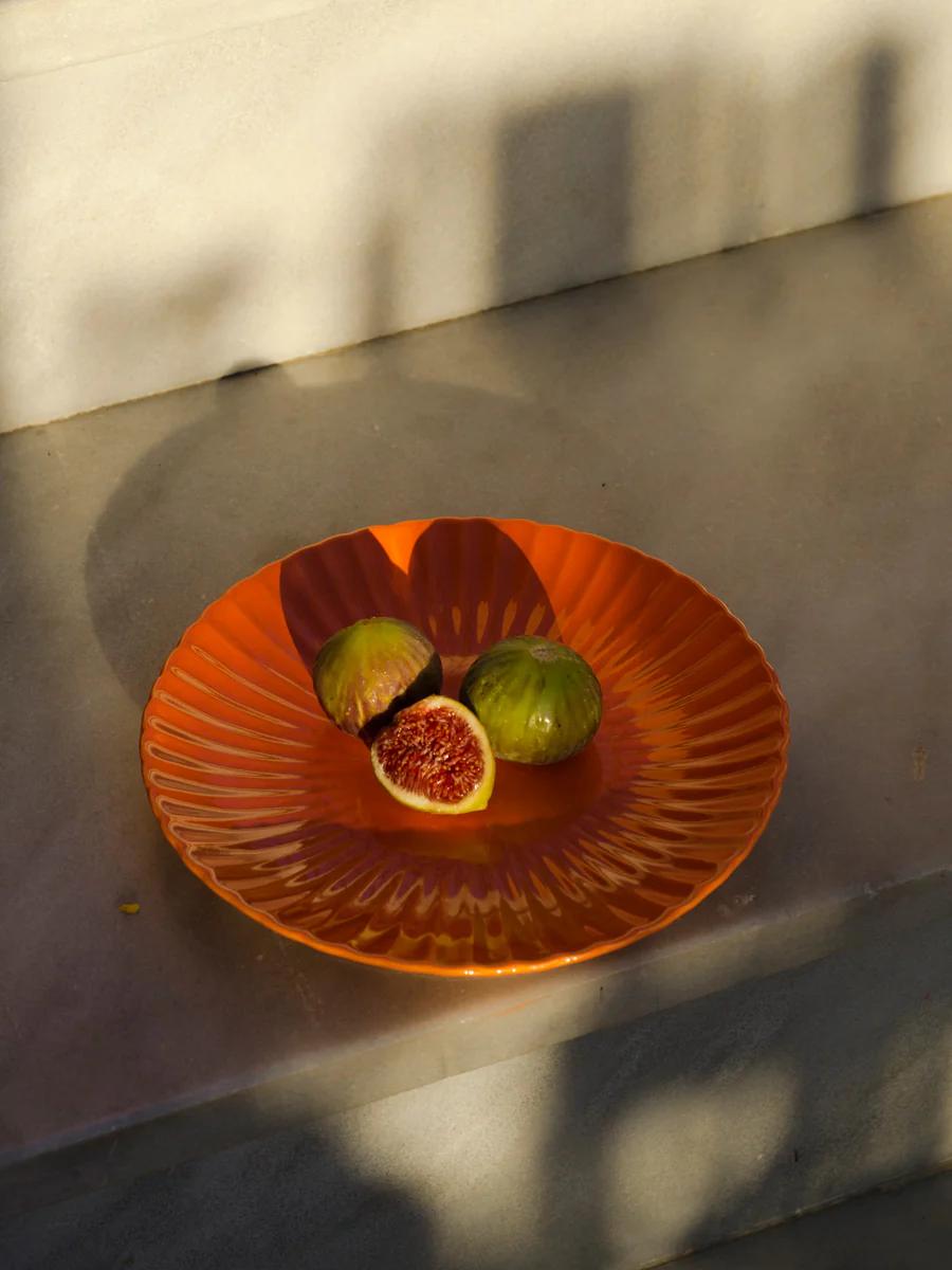 Orange fluted plate with three figs in sunlight. Two whole green figs, one cut open revealing red interior. Grey concrete surface casts soft shadows.