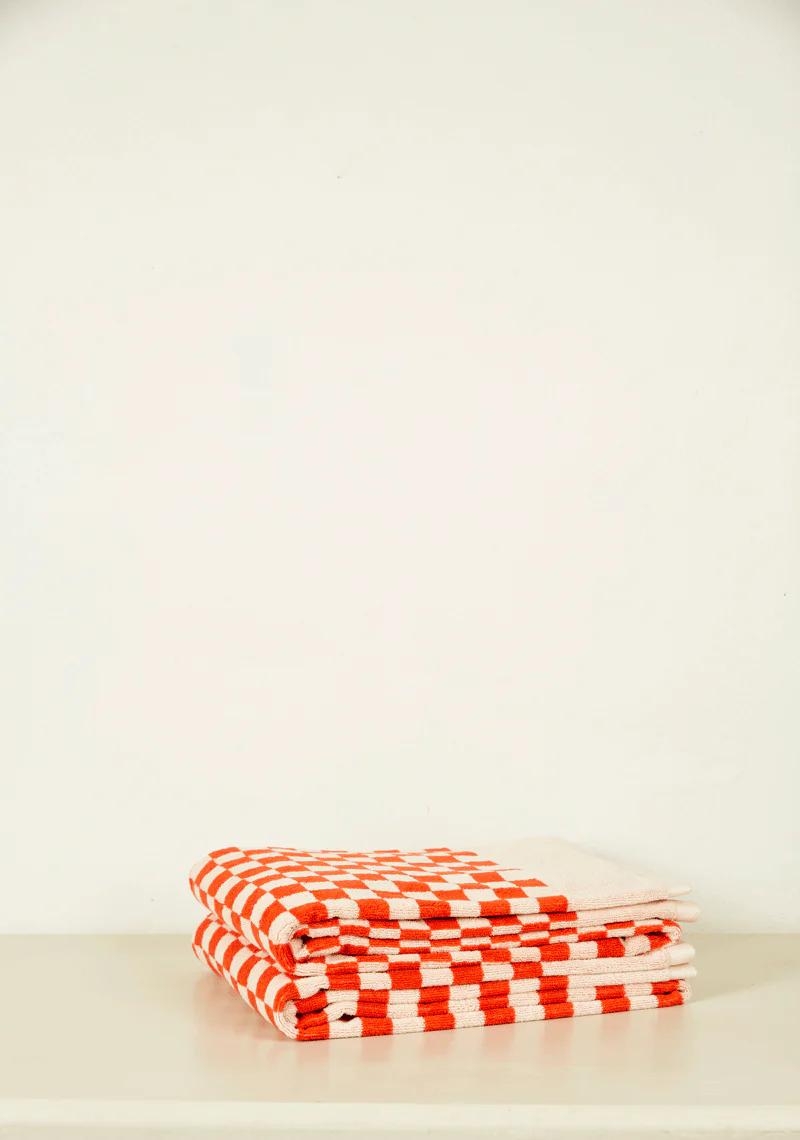 A folded stack of white and orange checkered organic cotton towels from Tessuti, neatly arranged on a white surface against a plain white wall.