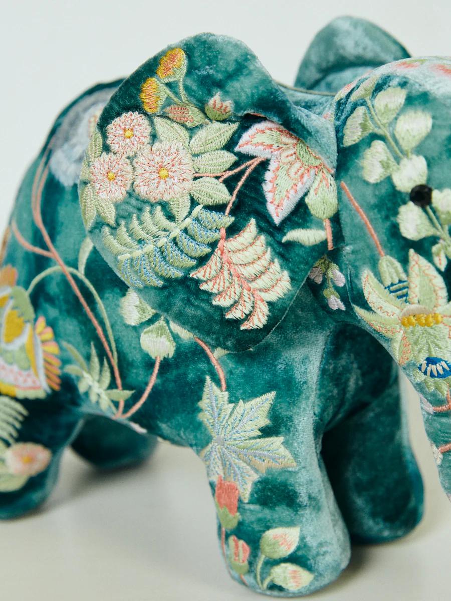 A plush elephant toy with teal velvet fabric stands on a light surface. Its body is covered in colourful floral embroidery featuring pink, yellow, green, and white stitching.