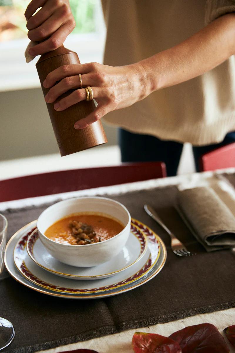 Hands season soup with a wooden pepper grinder. The table is set with plates, cutlery, napkin, and glass. A window is visible in the background.