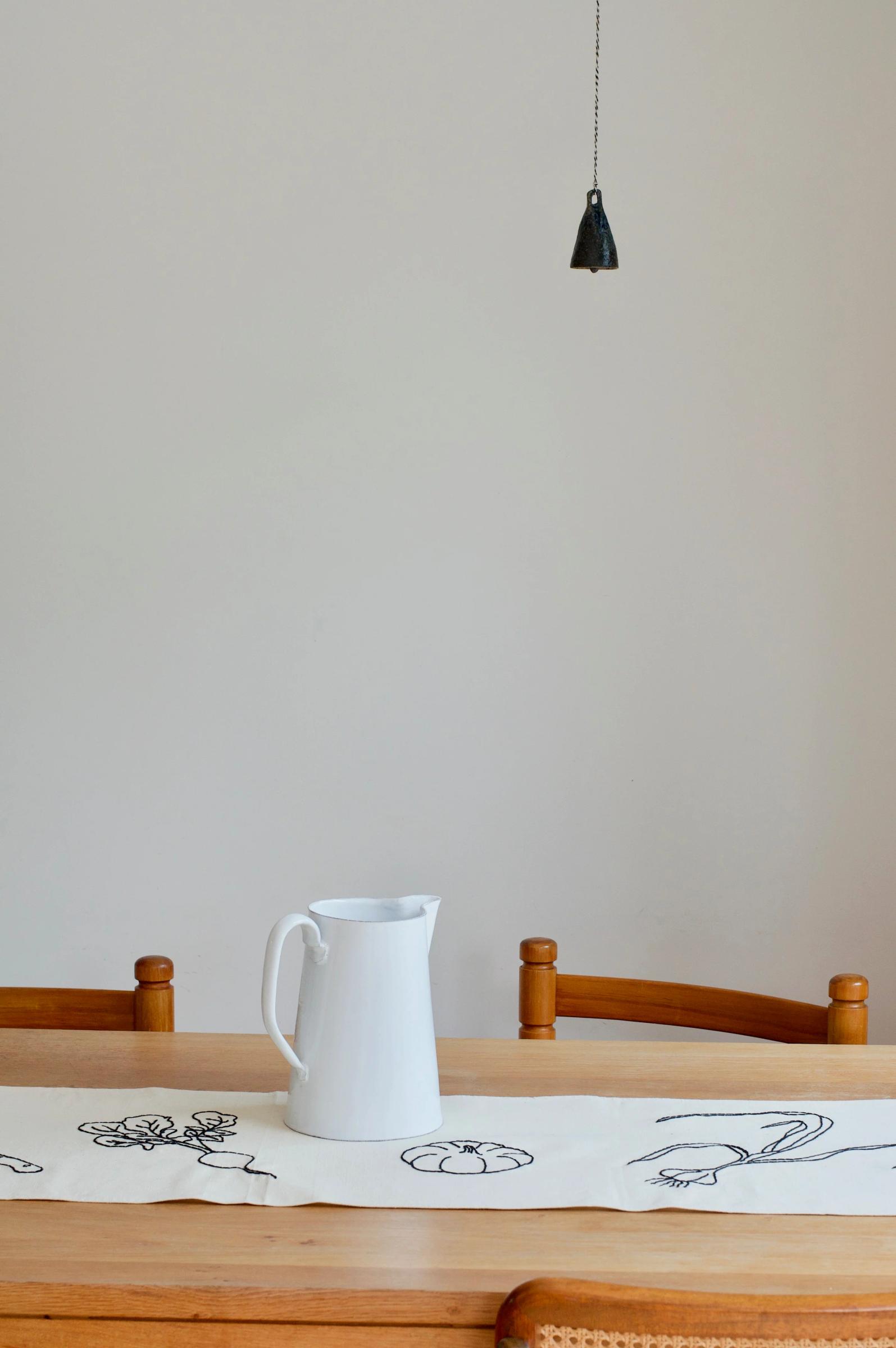 Wooden table and chairs, white ceramic pitcher on embroidered linens. Black pendant light with pull chain above. Beige wall background.