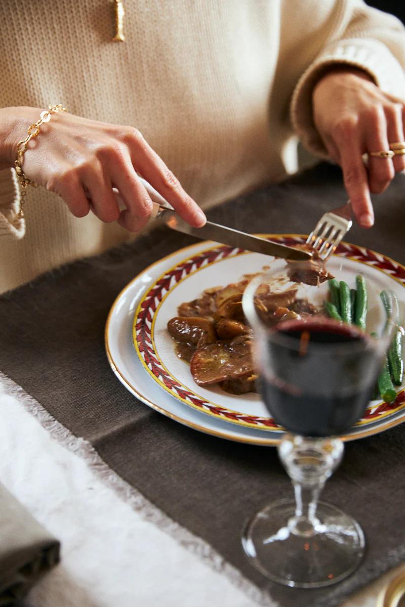 Person in beige sweater cuts into beef casserole with green beans. Red wine glass in foreground. Dark tablecloth.