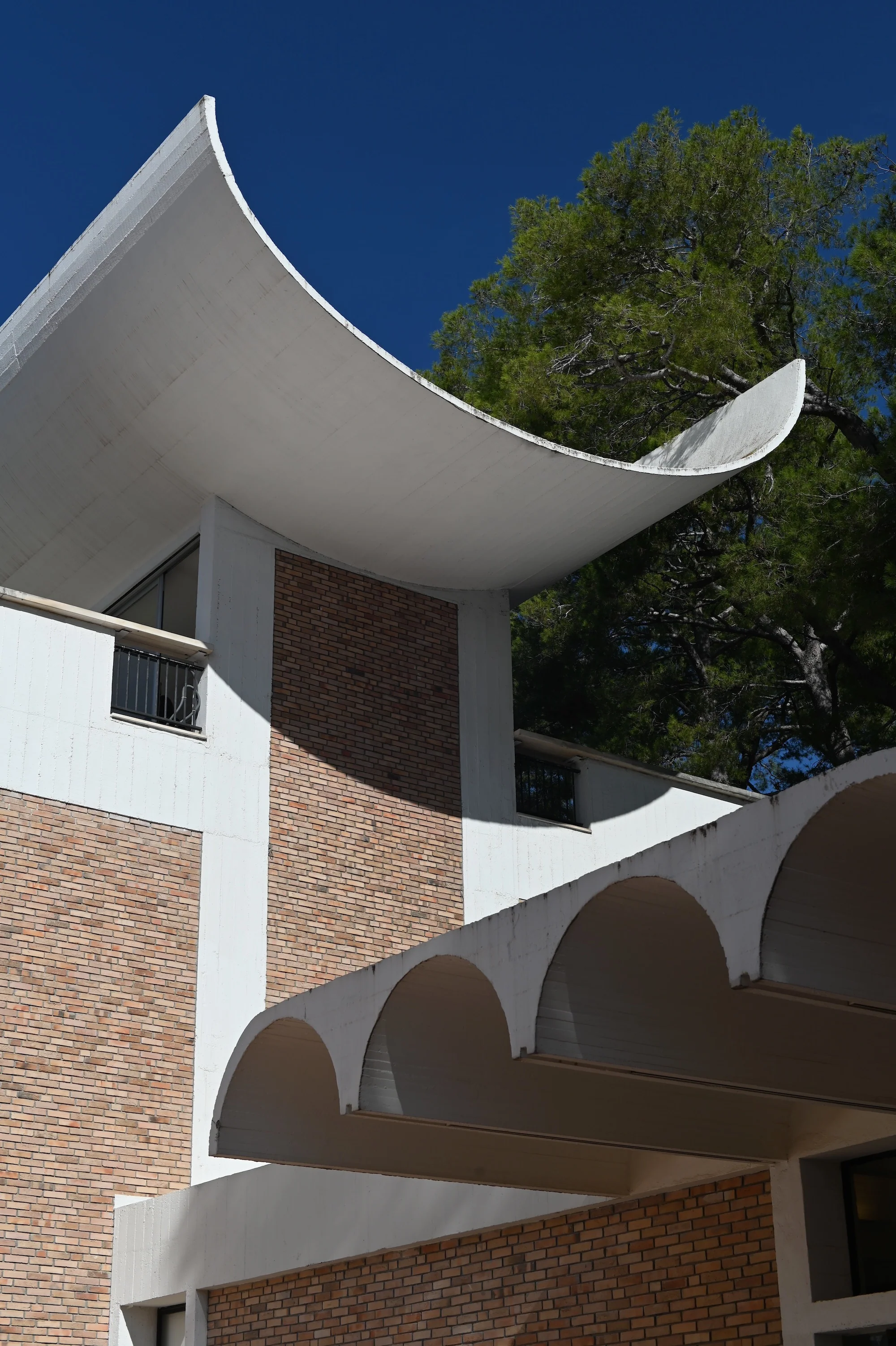 Modern building with curved roof. Brick and white concrete with arch motifs. Green backdrop, blue sky.