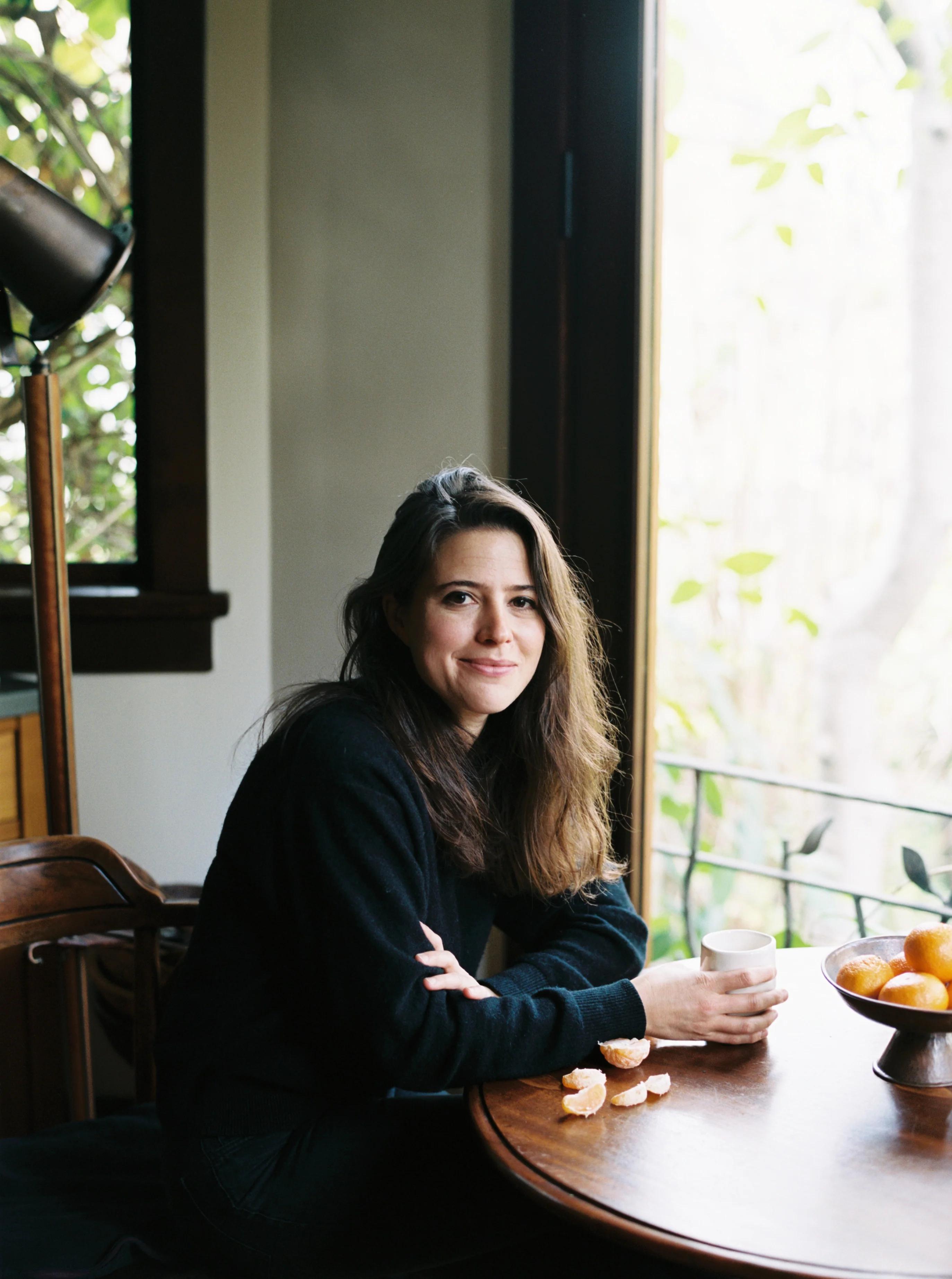 Woman with long brown hair at wooden table by window. Holding cup, smiling. Orange segments and bowl on table.