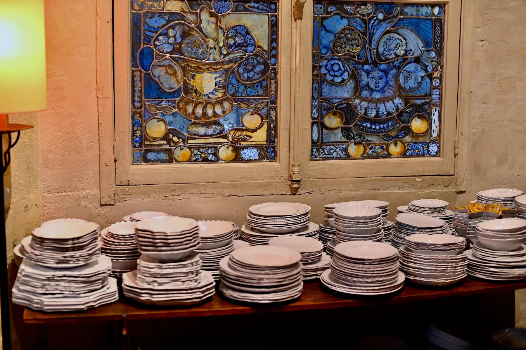 Wooden table stacked with white ceramic plates. Stained glass window with blue and gold designs behind.