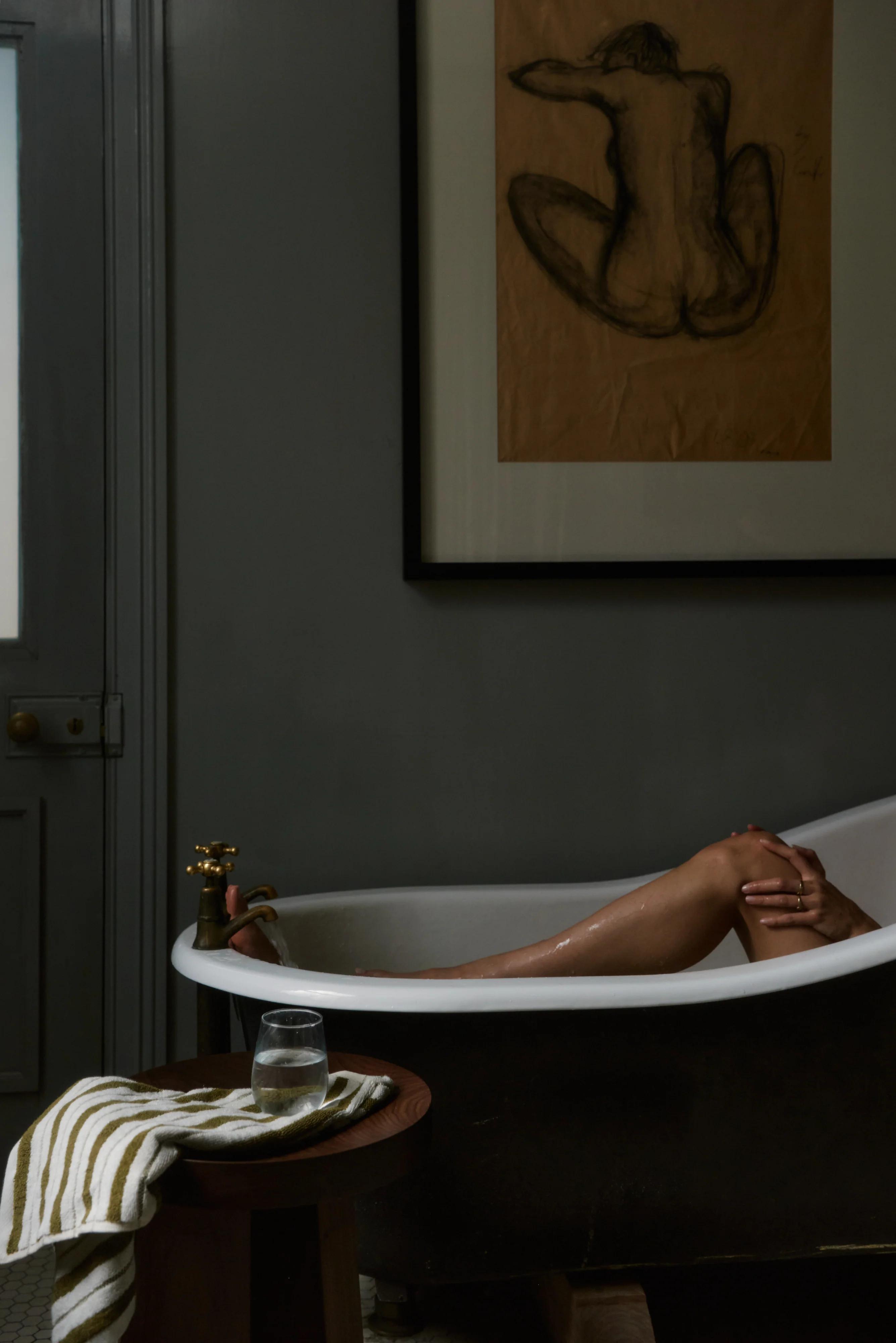 Person partially submerged in dimly lit bathtub. Framed sketch above. Striped towel and water glass on stool.