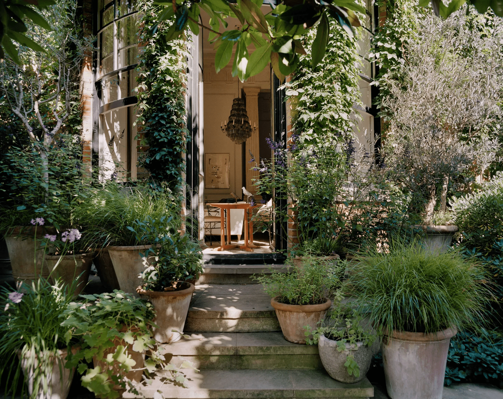 Lush garden leading to elegant doorway. Wooden table under chandelier inside.