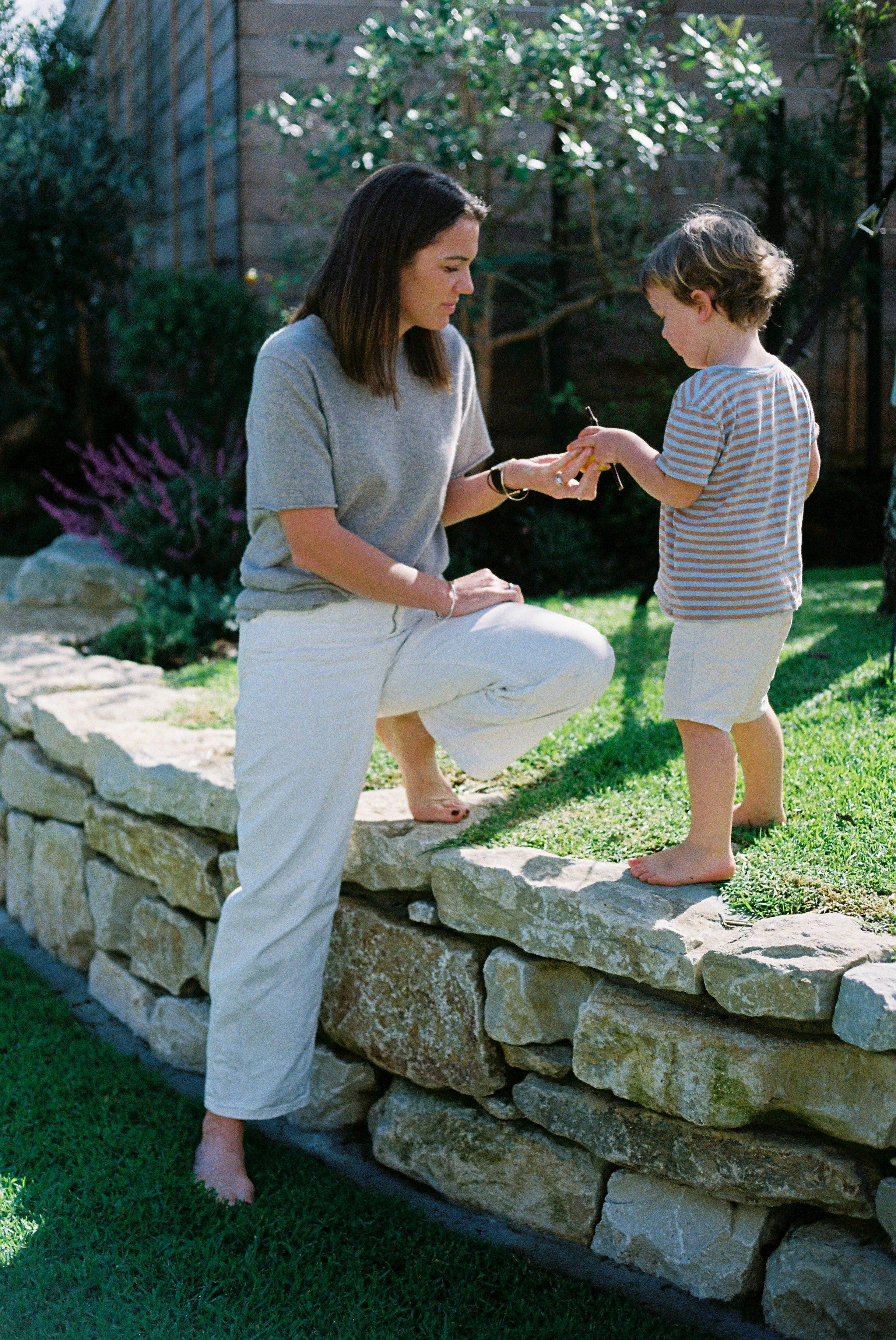 Woman in grey shirt and white pants helps child on low stone wall. Child in striped shirt and light shorts holds object. Garden setting.