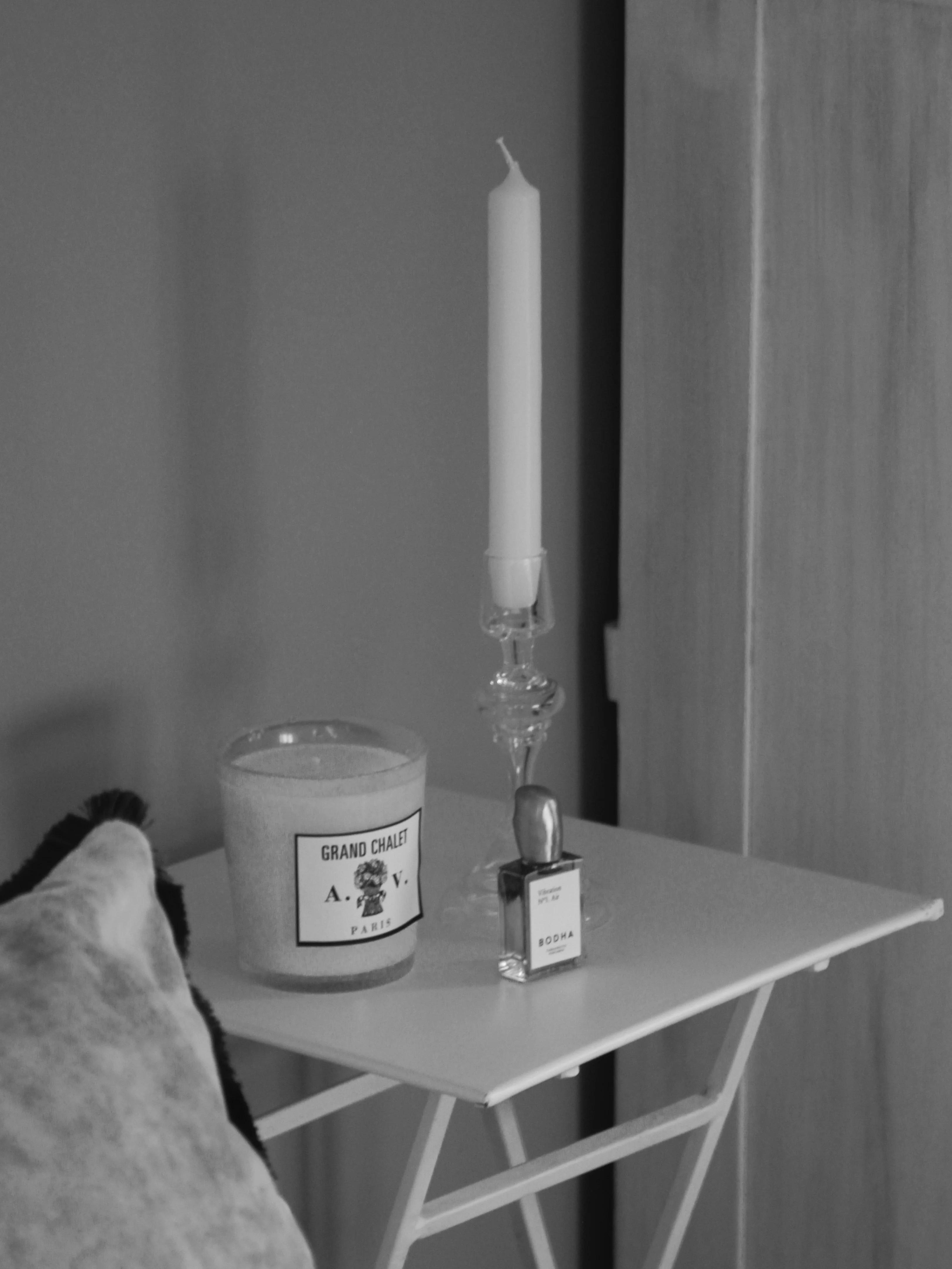 Black and white image of table with candle, perfume bottle, and glass candleholder. Chair with draped blanket visible, evoking intimate winter ritual setting.