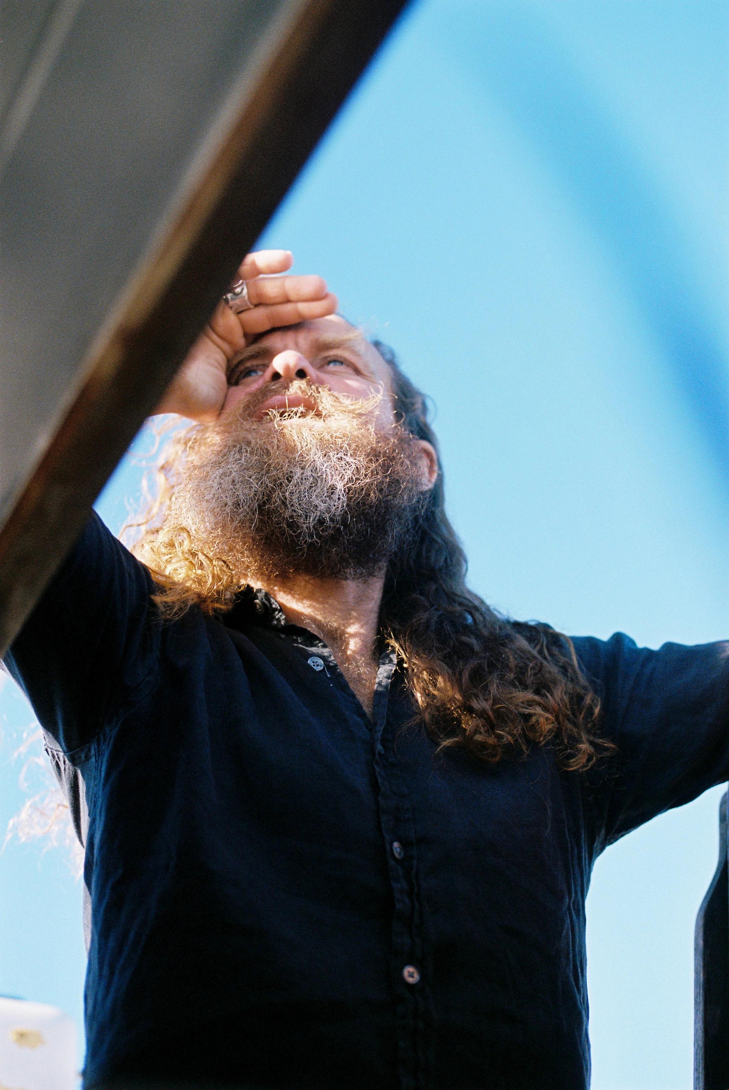 Person with long hair and beard looking up, shading eyes. Dark shirt, blue sky background.