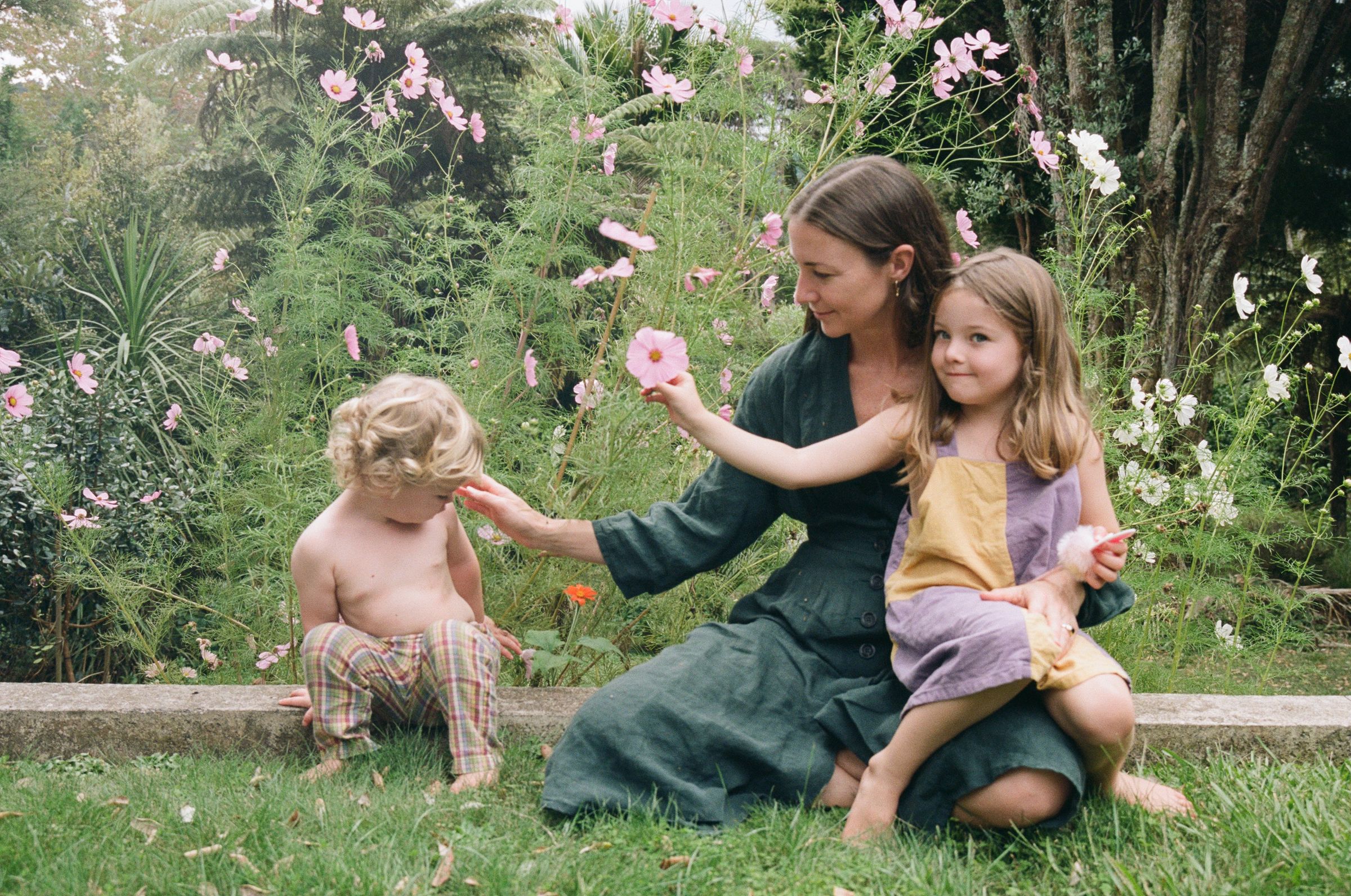 Woman on grass with two children. Holding one, touching face of other. Surrounded by greenery and pink flowers.