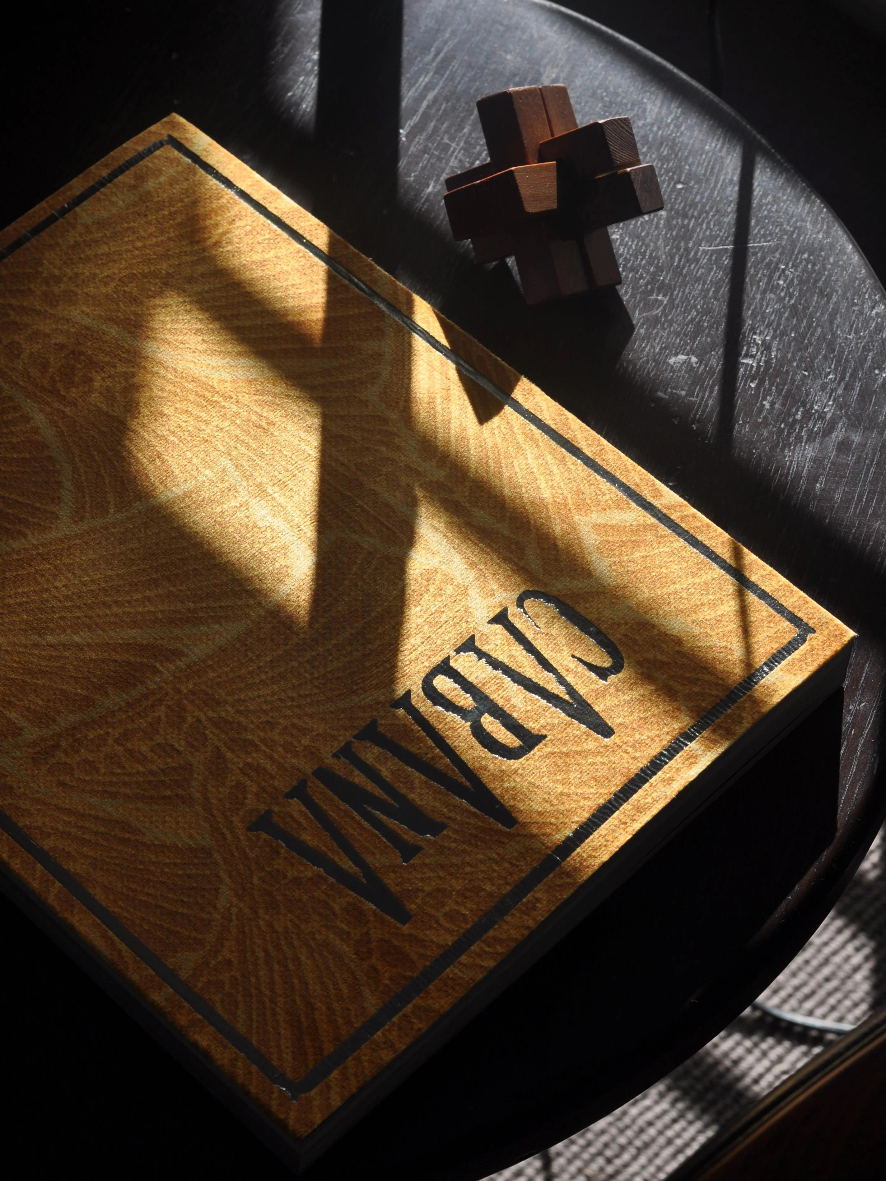 Yellow "CABANA" book on dark wooden table. Sunlight casting cross-shaped shadow. Small wooden puzzle nearby.