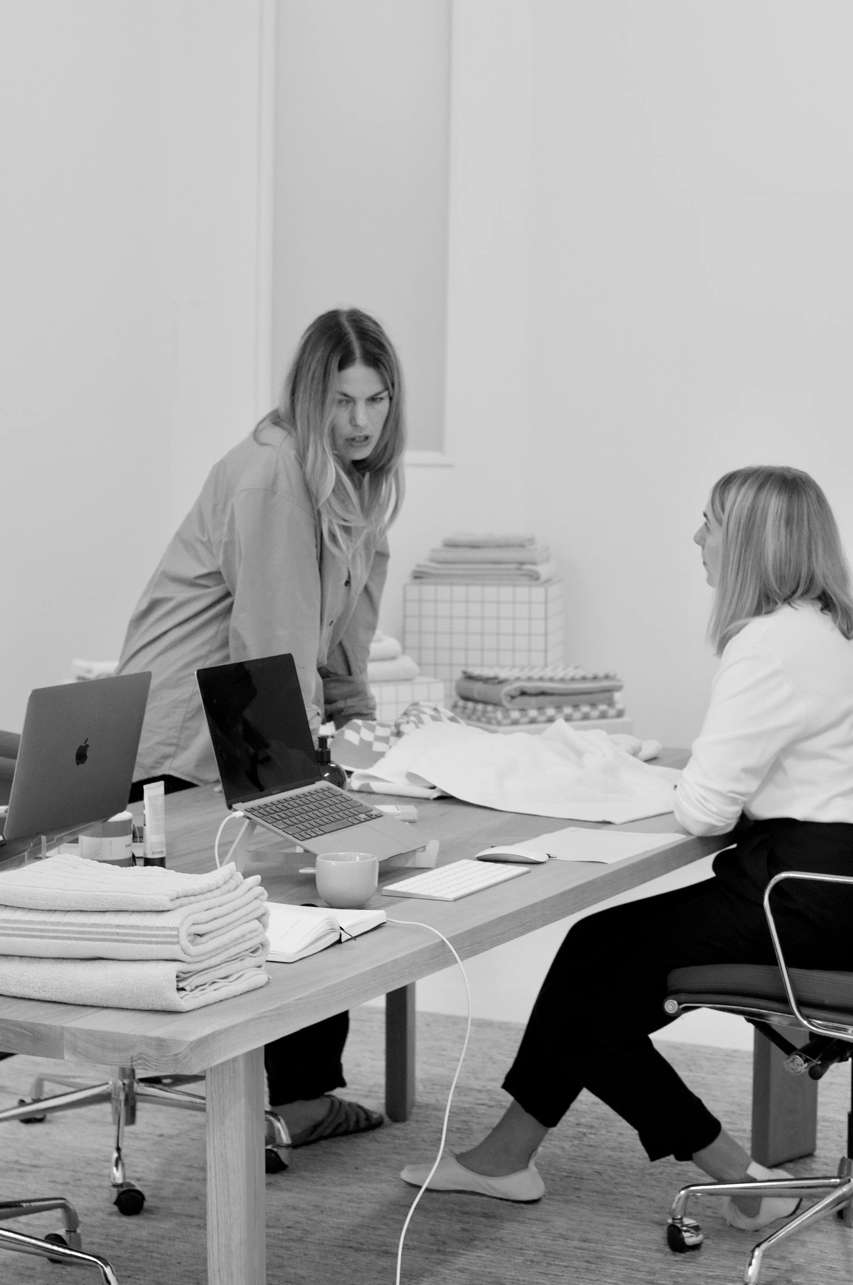 Black and white photo of two people discussing in modern office. Laptops, fabric samples on table. Folded fabric stacks visible.