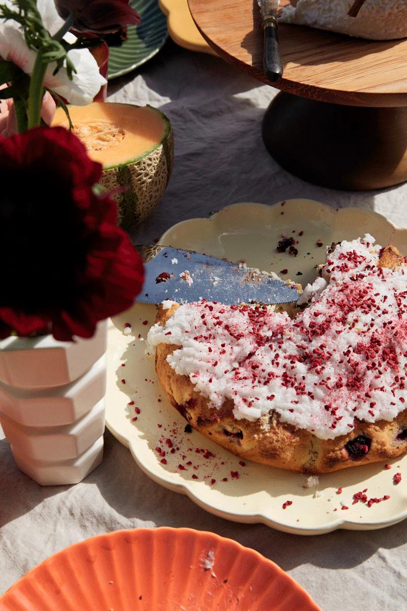 Partially sliced cake with white and pink frosting on scalloped plate. Background shows cantaloupe, wooden cake stand, and flowers. Light tablecloth showcases easy baking.