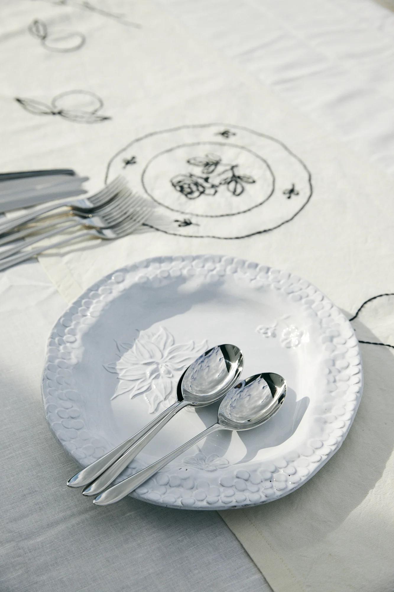 White embossed plate on floral embroidered tablecloth. Teaspoons, forks, and knives neatly arranged beside.