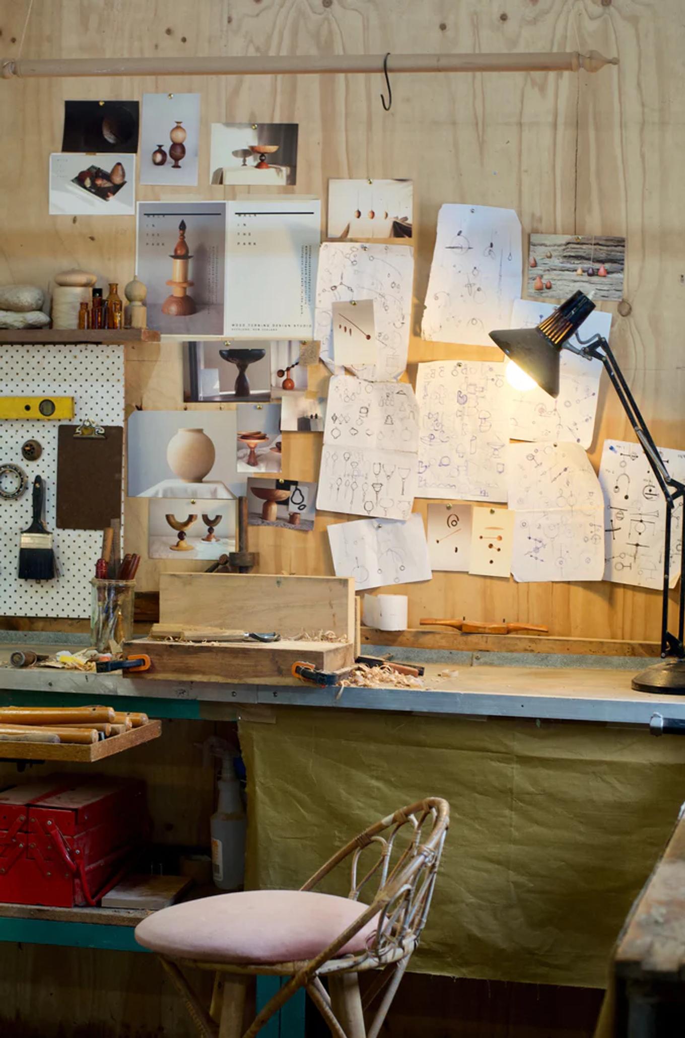 Cluttered woodworking studio with bench, tools, and sketches. Lamp illuminates workspace with cushioned chair. Atmosphere evokes NZ wood craft ambiance.